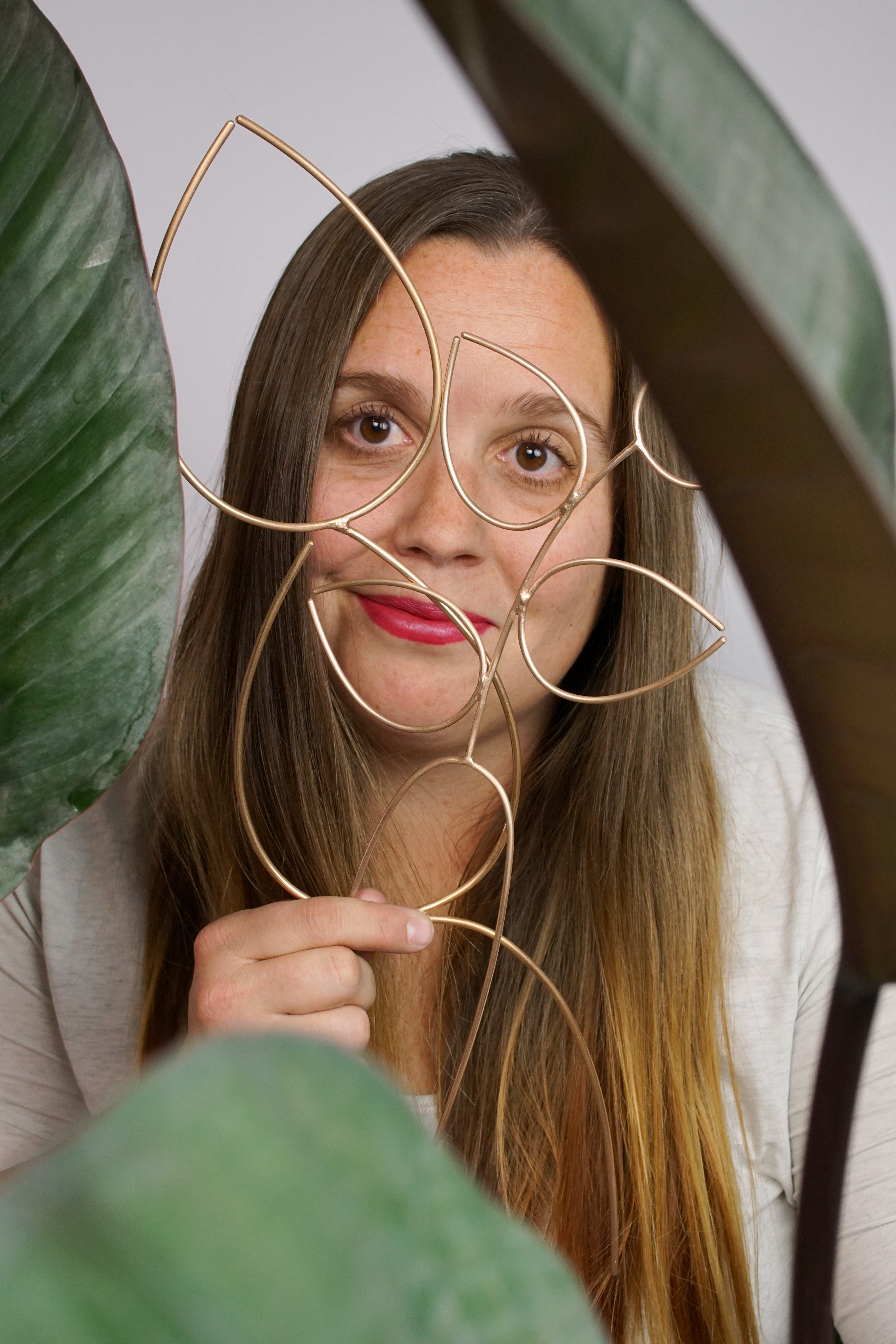 thoughts on scale: artist, designer, and metalsmith megan auman holds a pair of bronze plant stakes while sitting behind a philodendron rojo congo