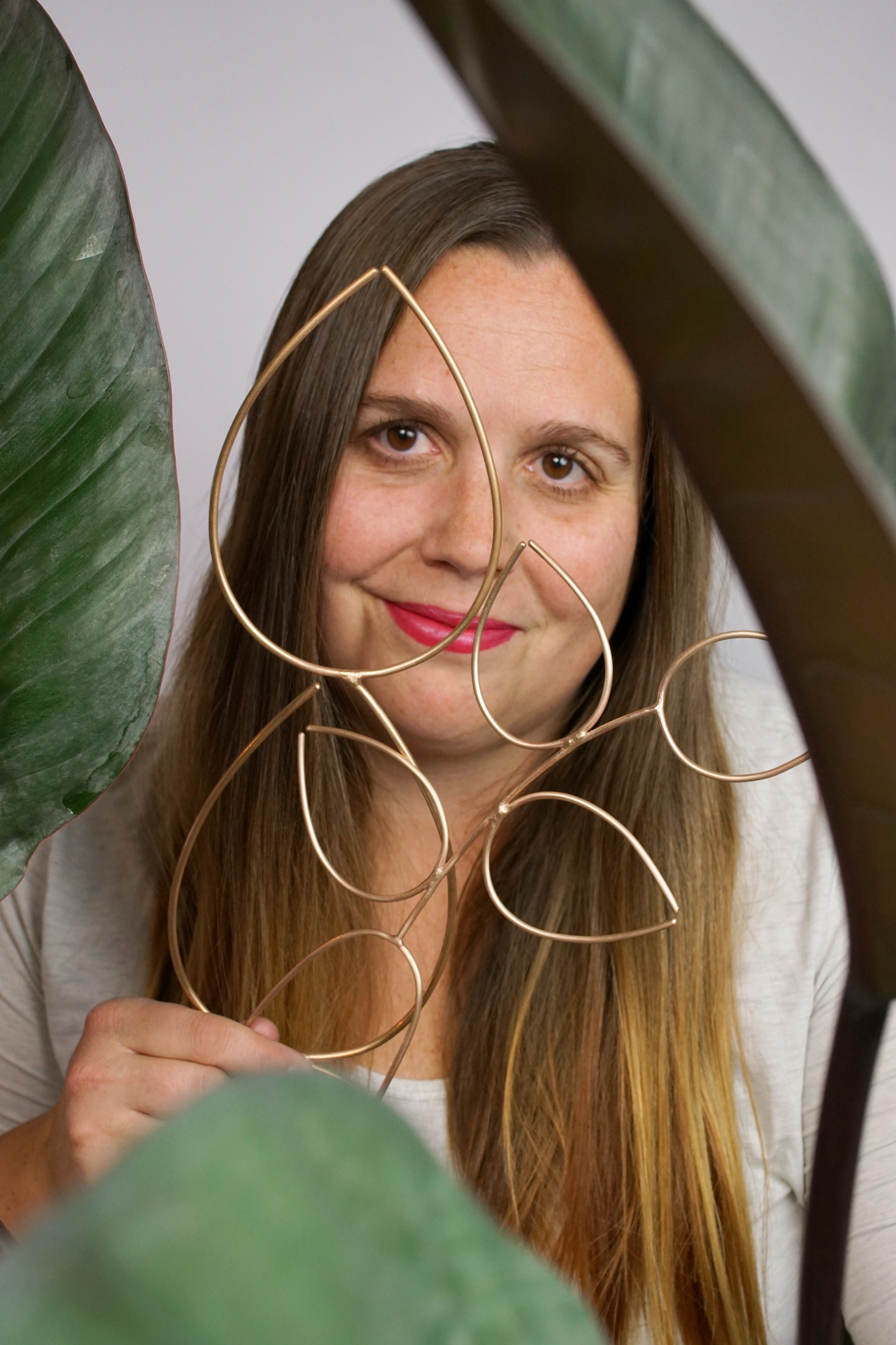 thoughts on scale: artist, designer, and metalsmith megan auman holds a pair of bronze plant stakes while sitting behind a philodendron rojo congo