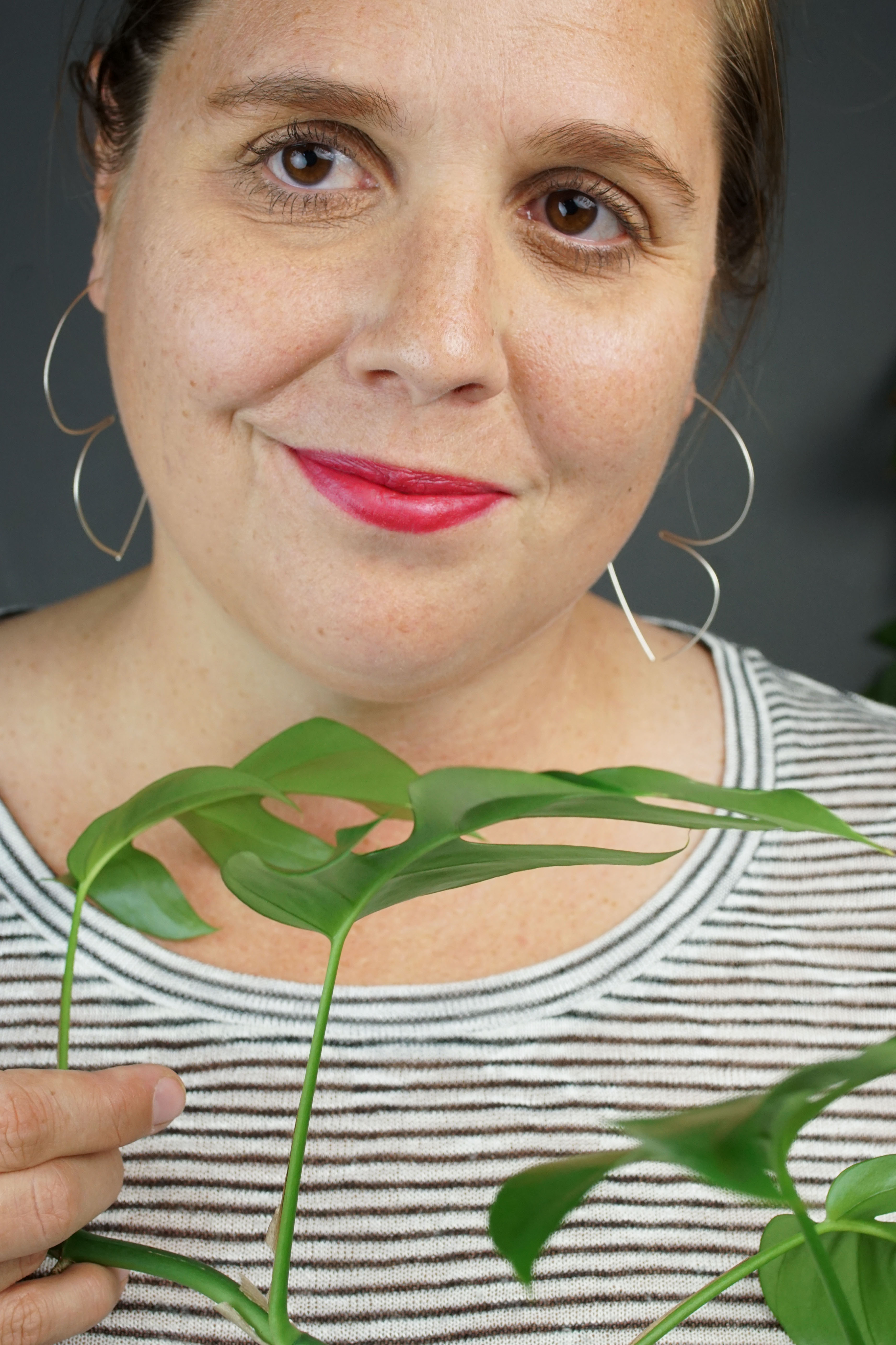 woman with red lipstick wearing silver hoop threader earrings and a striped shirt and hiding behind a plant