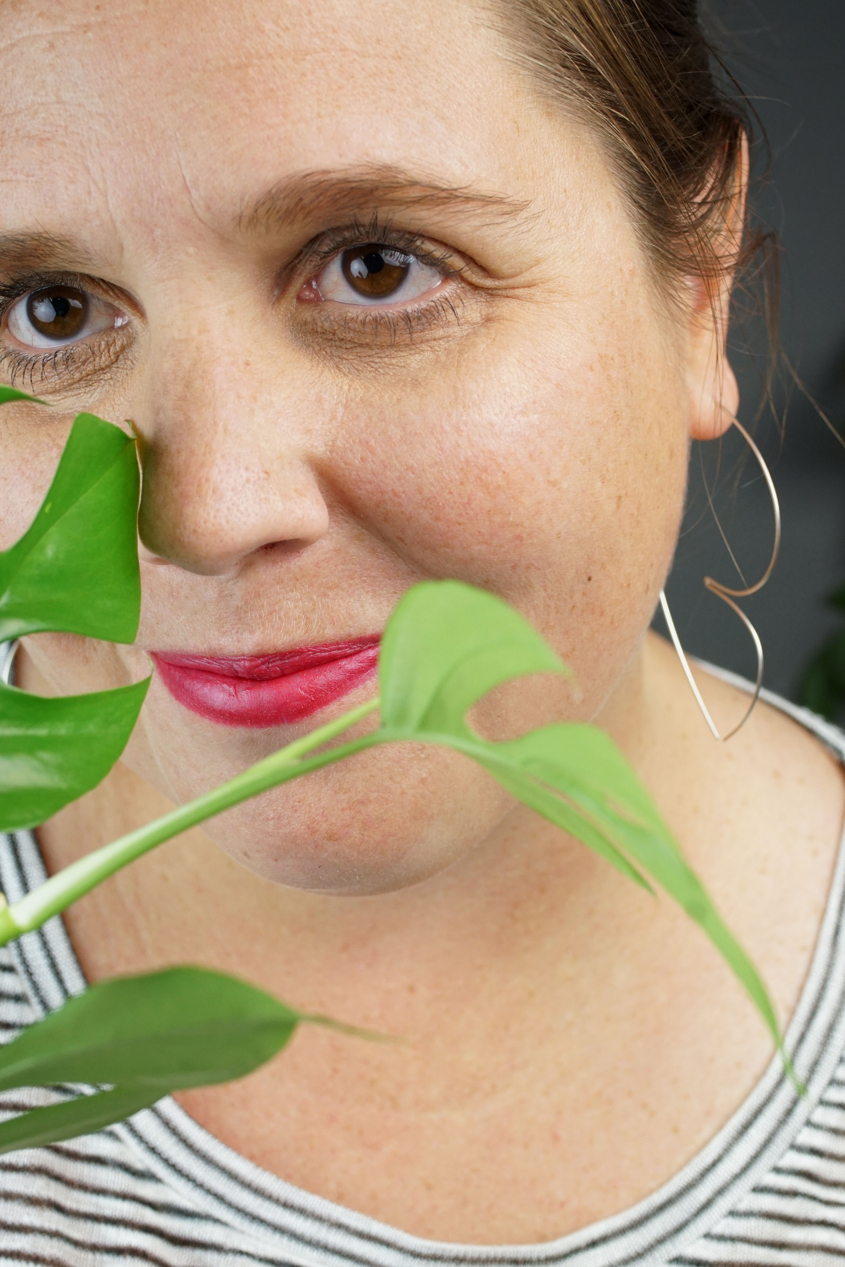 woman with red lipstick wearing silver hoop threader earrings and a striped shirt and hiding behind a plant