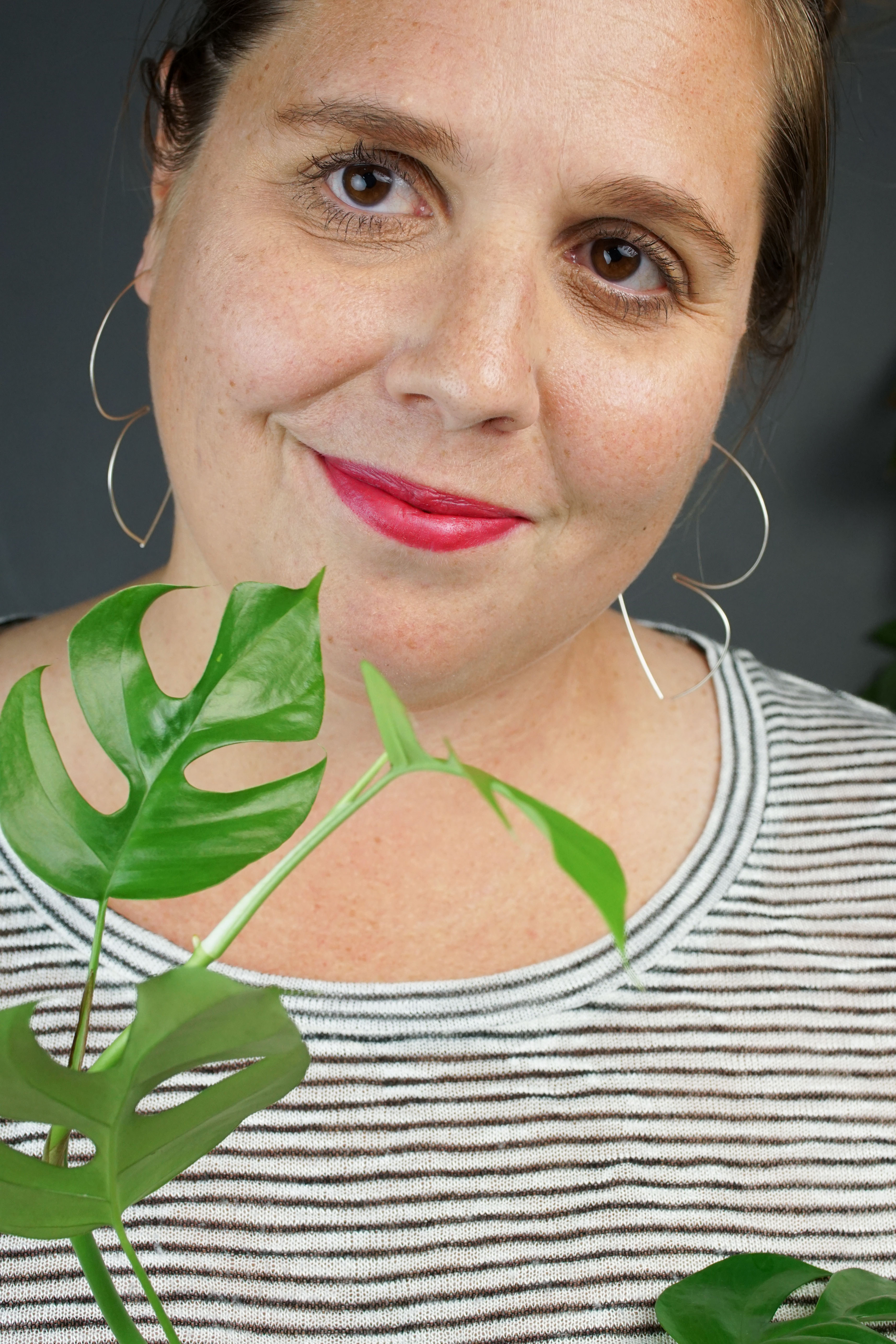 woman with red lipstick wearing silver hoop threader earrings and a striped shirt and hiding behind a plant