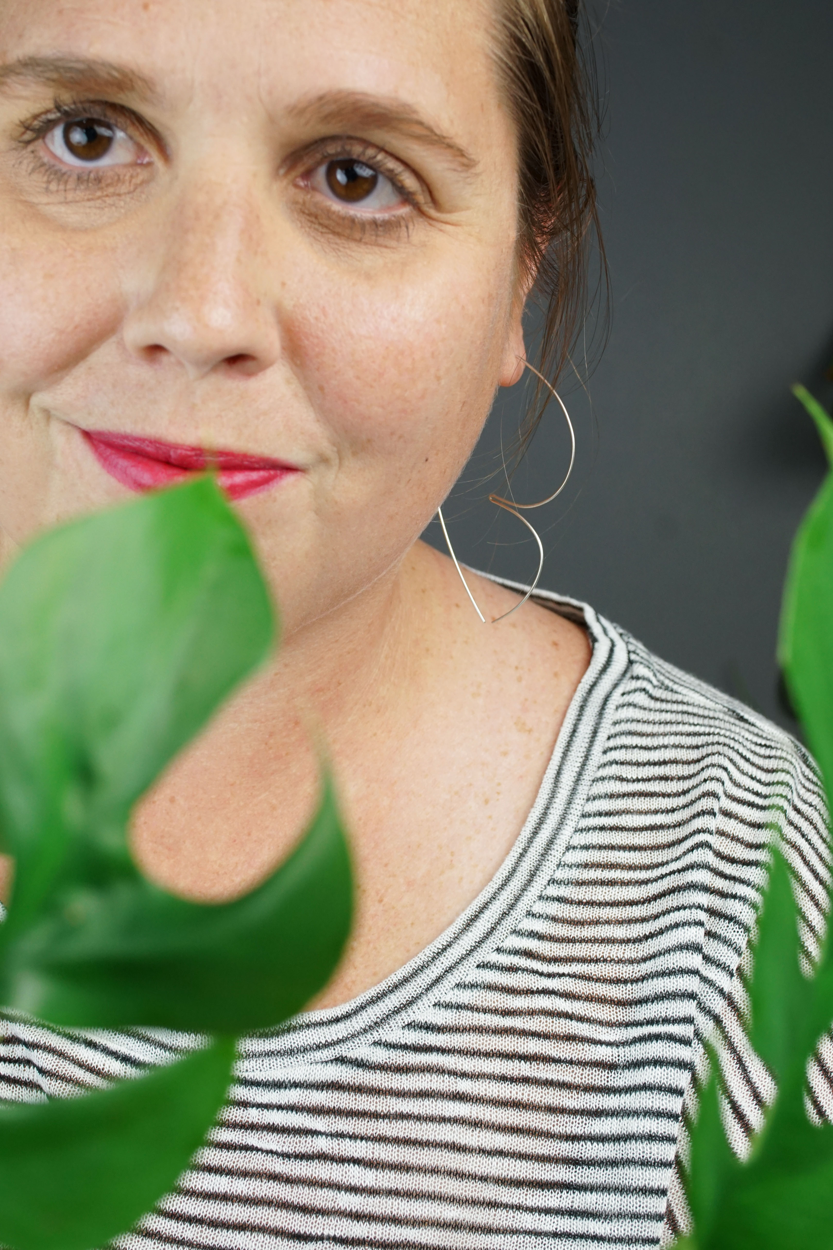 woman with red lipstick wearing silver hoop threader earrings and a striped shirt and hiding behind a plant