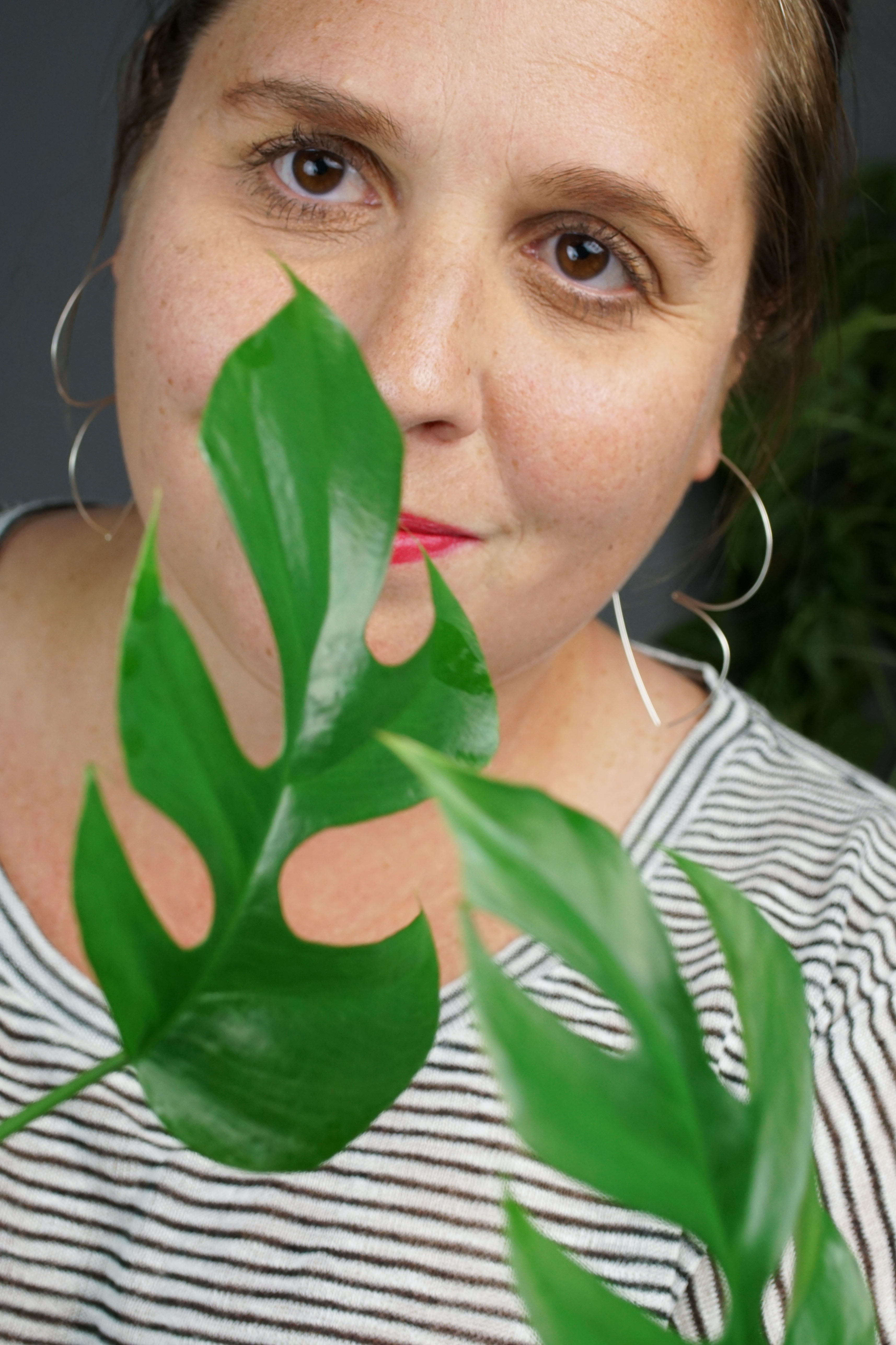 woman with red lipstick wearing silver hoop threader earrings and a striped shirt and hiding behind a plant