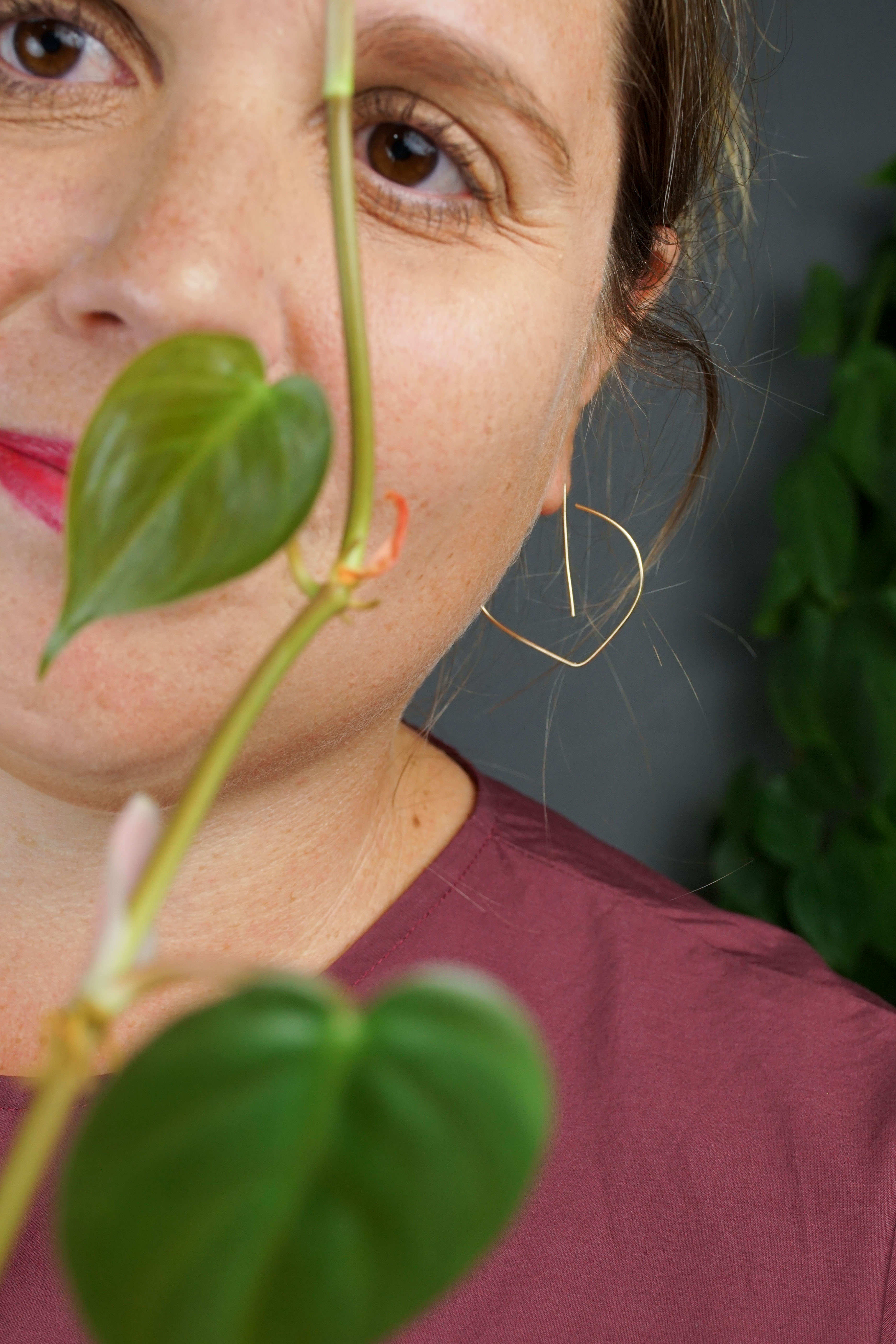 woman wearing burgundy dress and gold threader hoop earrings and standing in front of a heart-leaf philodendron