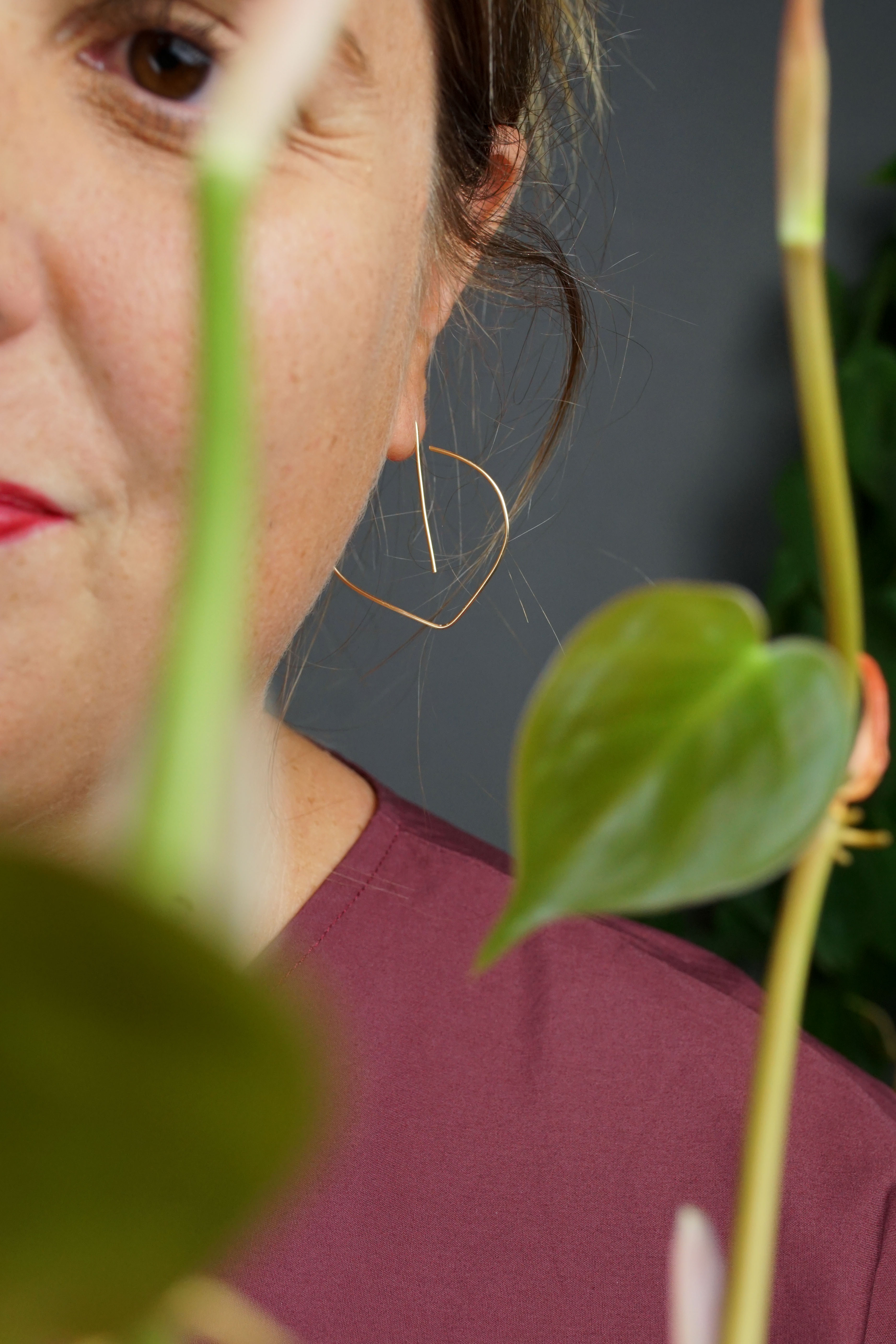 woman wearing burgundy dress and gold threader hoop earrings and standing in front of a heart-leaf philodendron