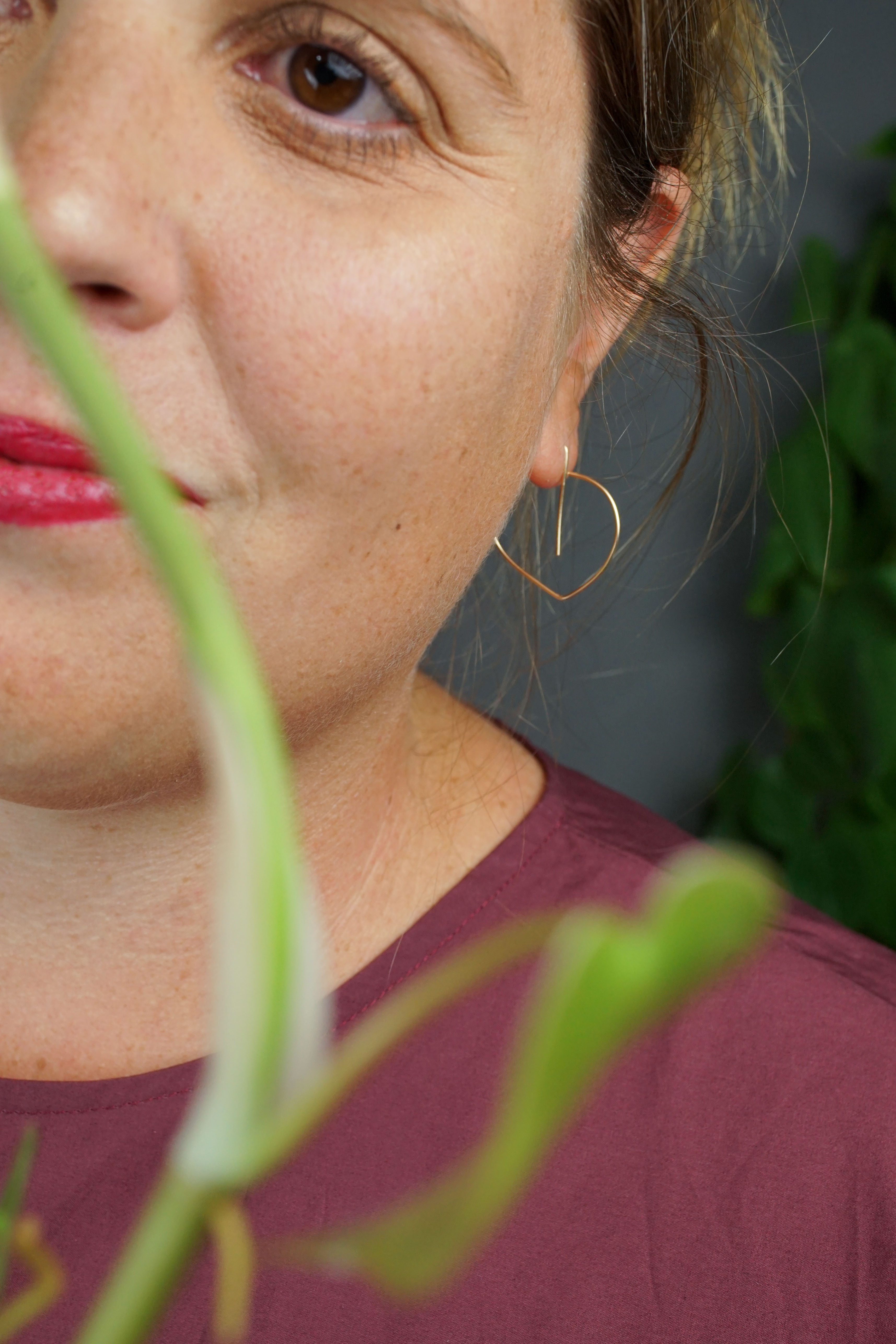 woman wearing burgundy dress and gold threader hoop earrings and standing in front of a heart-leaf philodendron