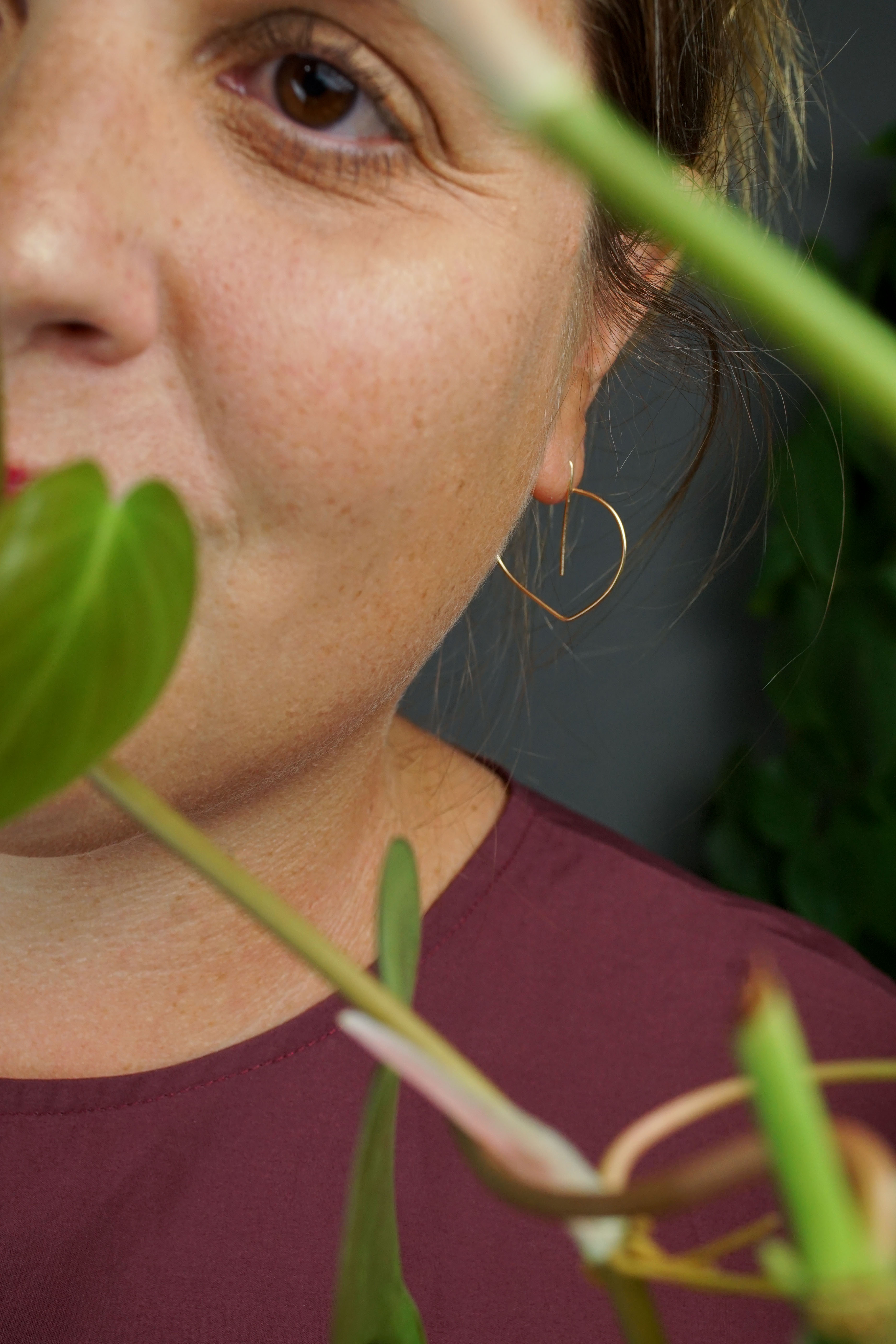 woman wearing burgundy dress and gold threader hoop earrings and standing in front of a heart-leaf philodendron