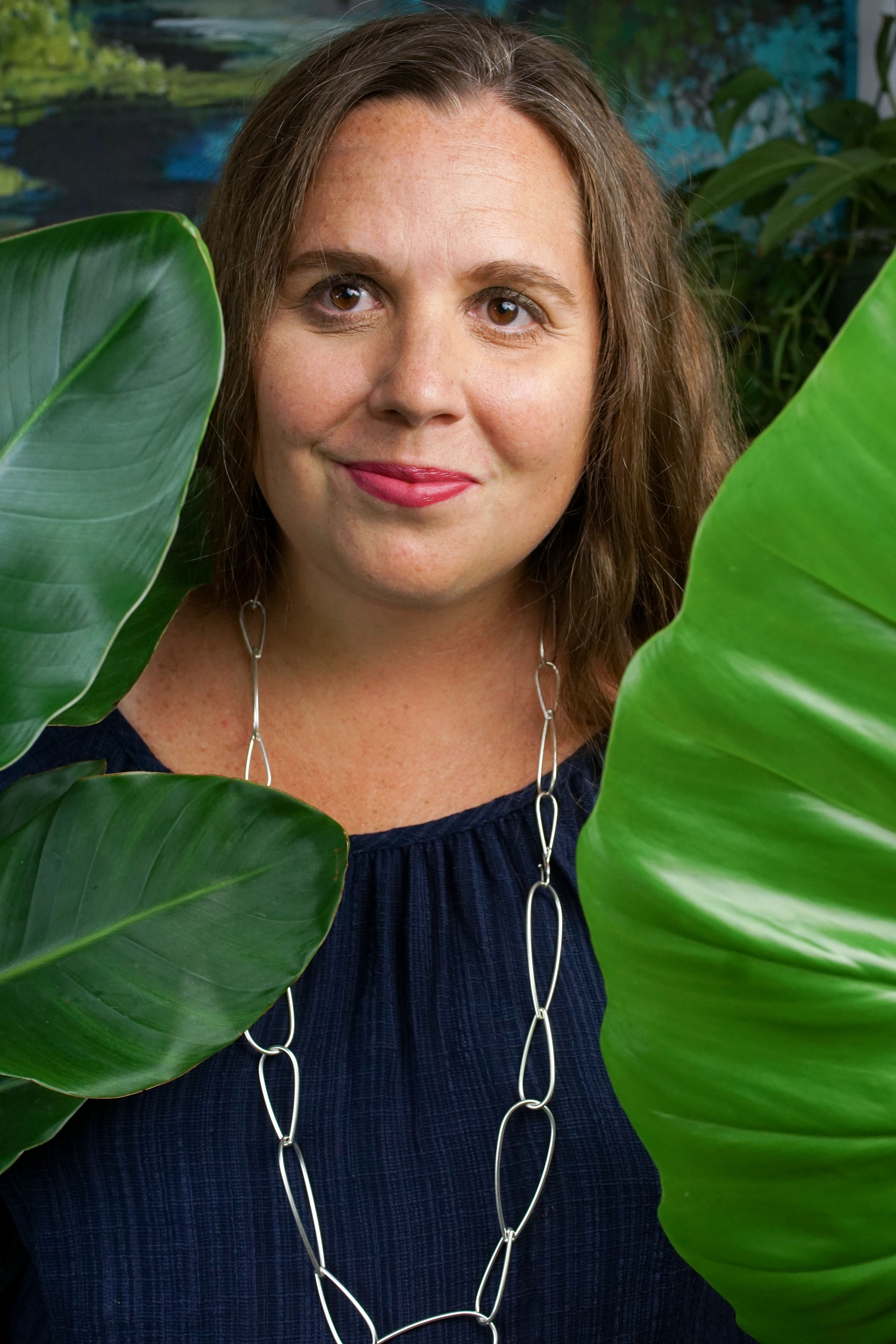 self portrait of woman wearing a blue dress and long silver chain necklace standing behind a philodendron giganteum and a bird of paradise house plants