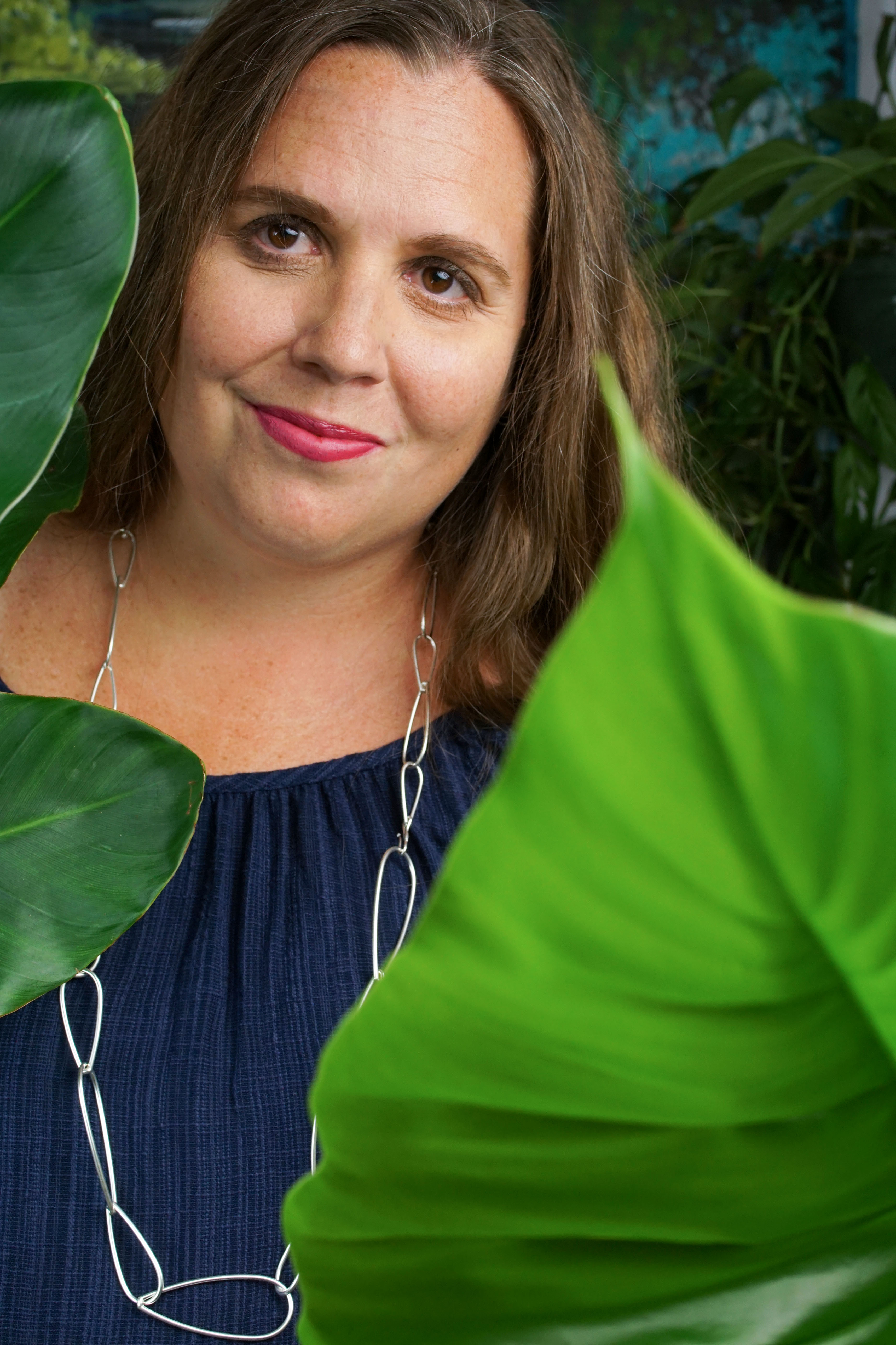 self portrait of woman wearing a blue dress and long silver chain necklace standing behind a philodendron giganteum and a bird of paradise house plants