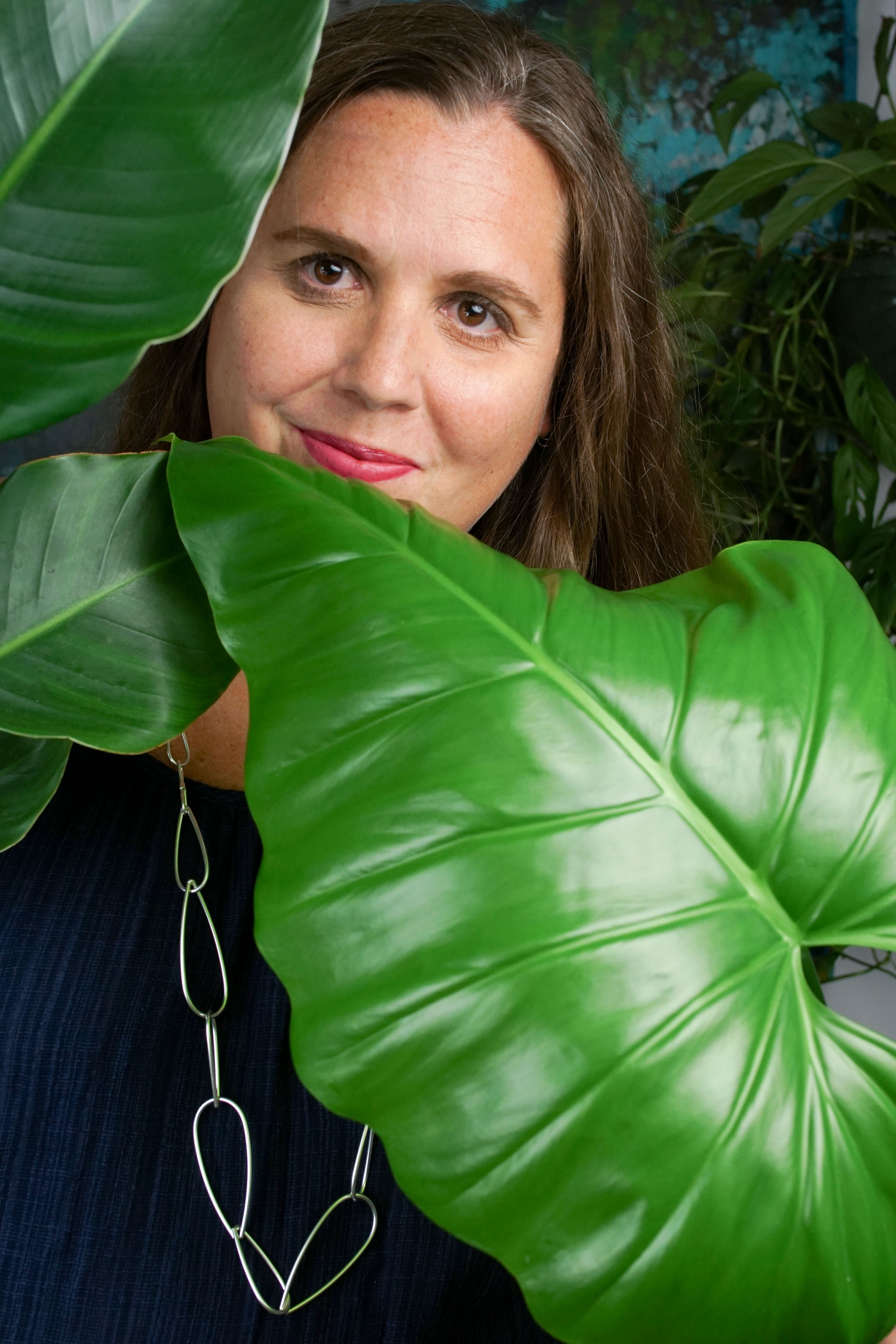 self portrait of woman wearing a blue dress and long silver chain necklace standing behind a philodendron giganteum and a bird of paradise house plants