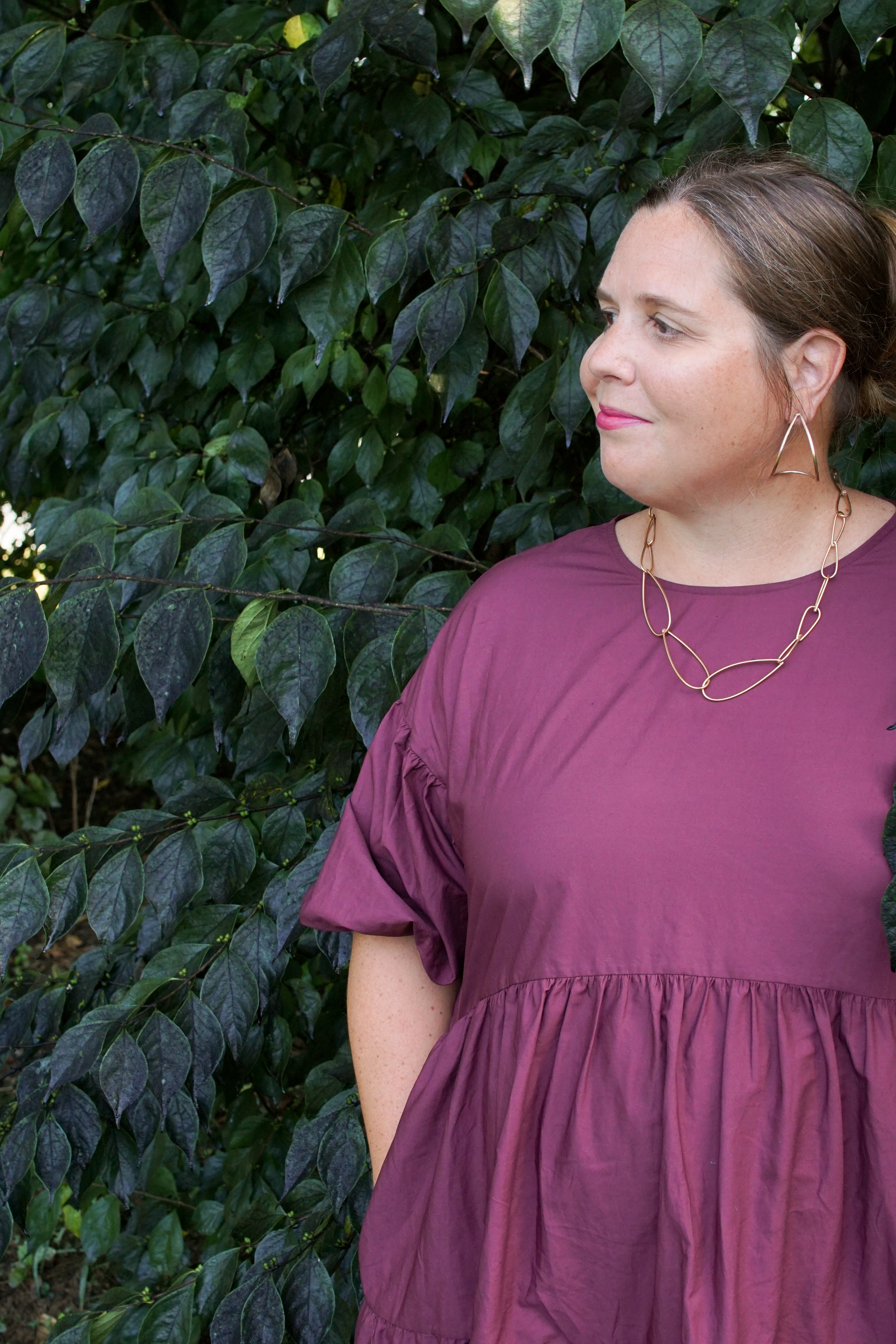 woman wearing burgundy dress and bronze chain necklace standing with a bush