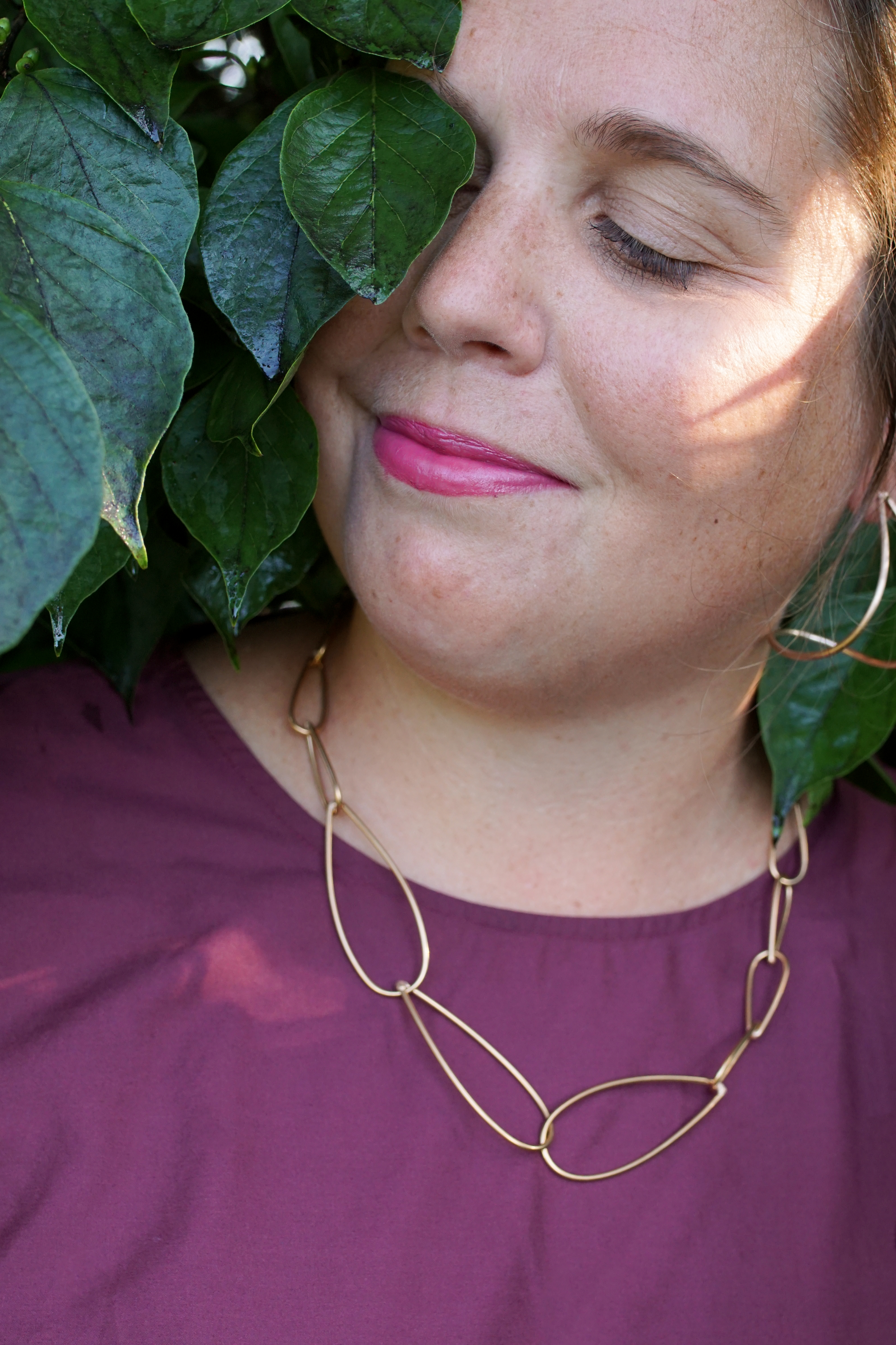 woman wearing burgundy dress and bronze chain necklace standing with a bush