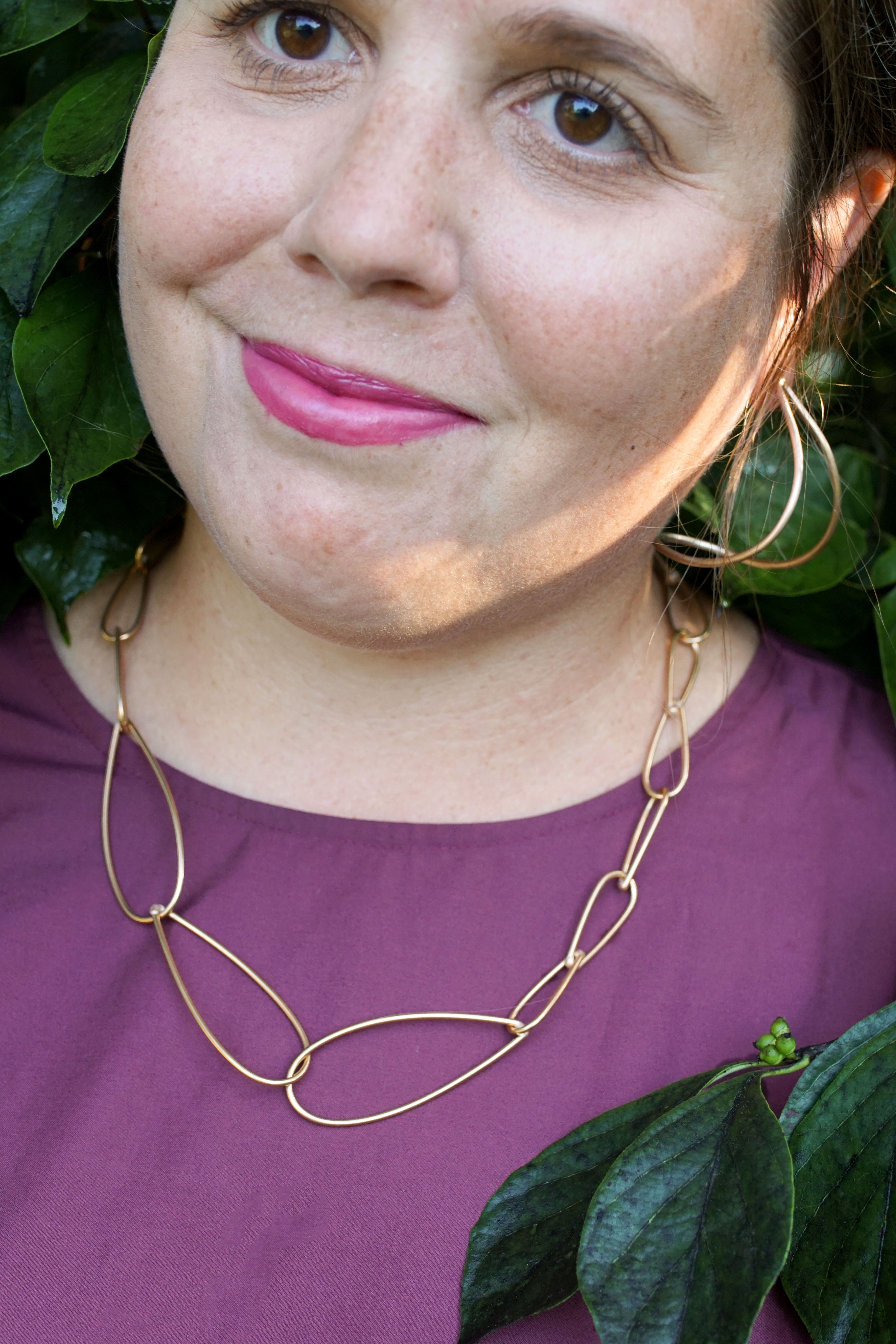 woman wearing burgundy dress and bronze chain necklace standing with a bush