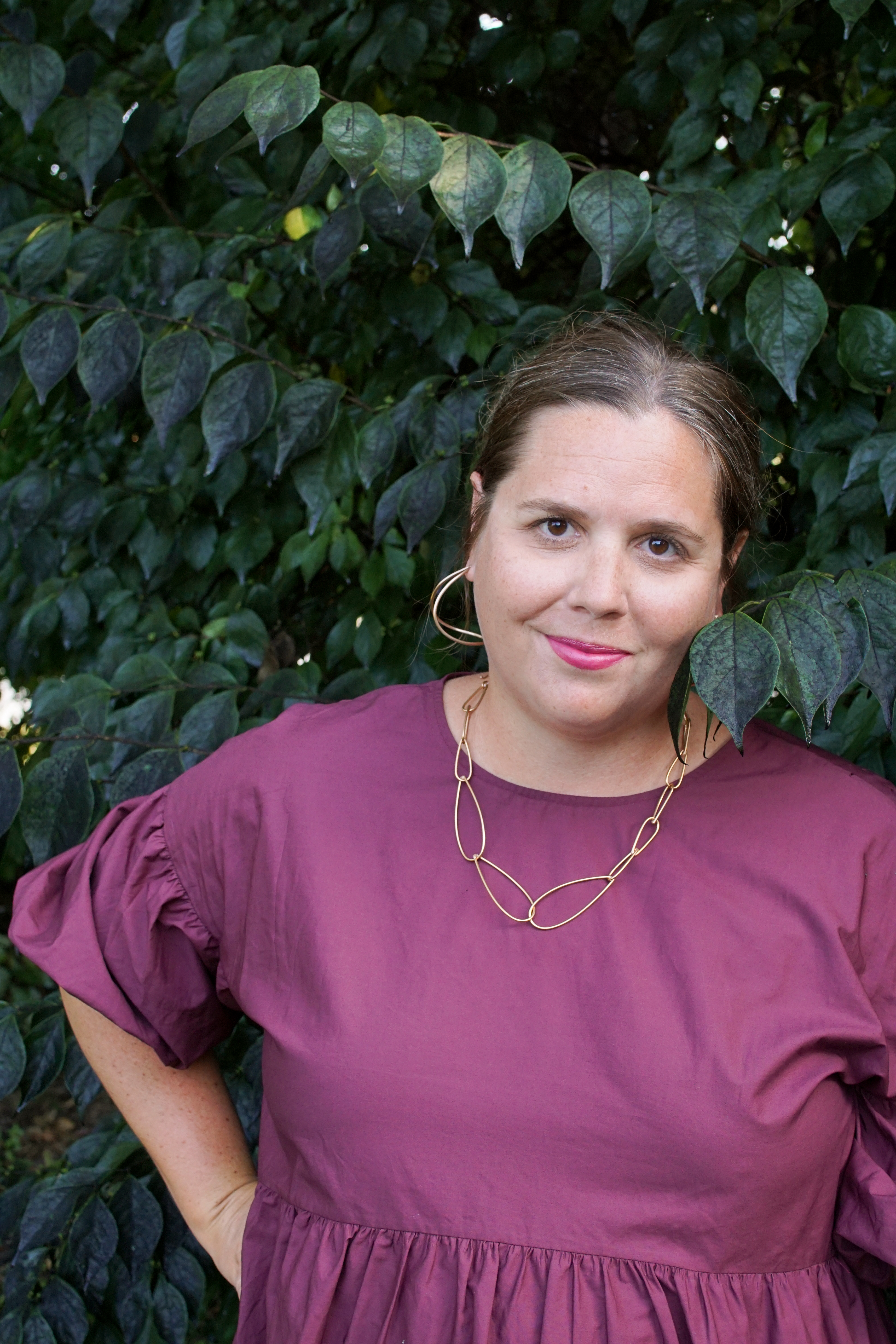 woman wearing burgundy dress and bronze chain necklace standing with a bush