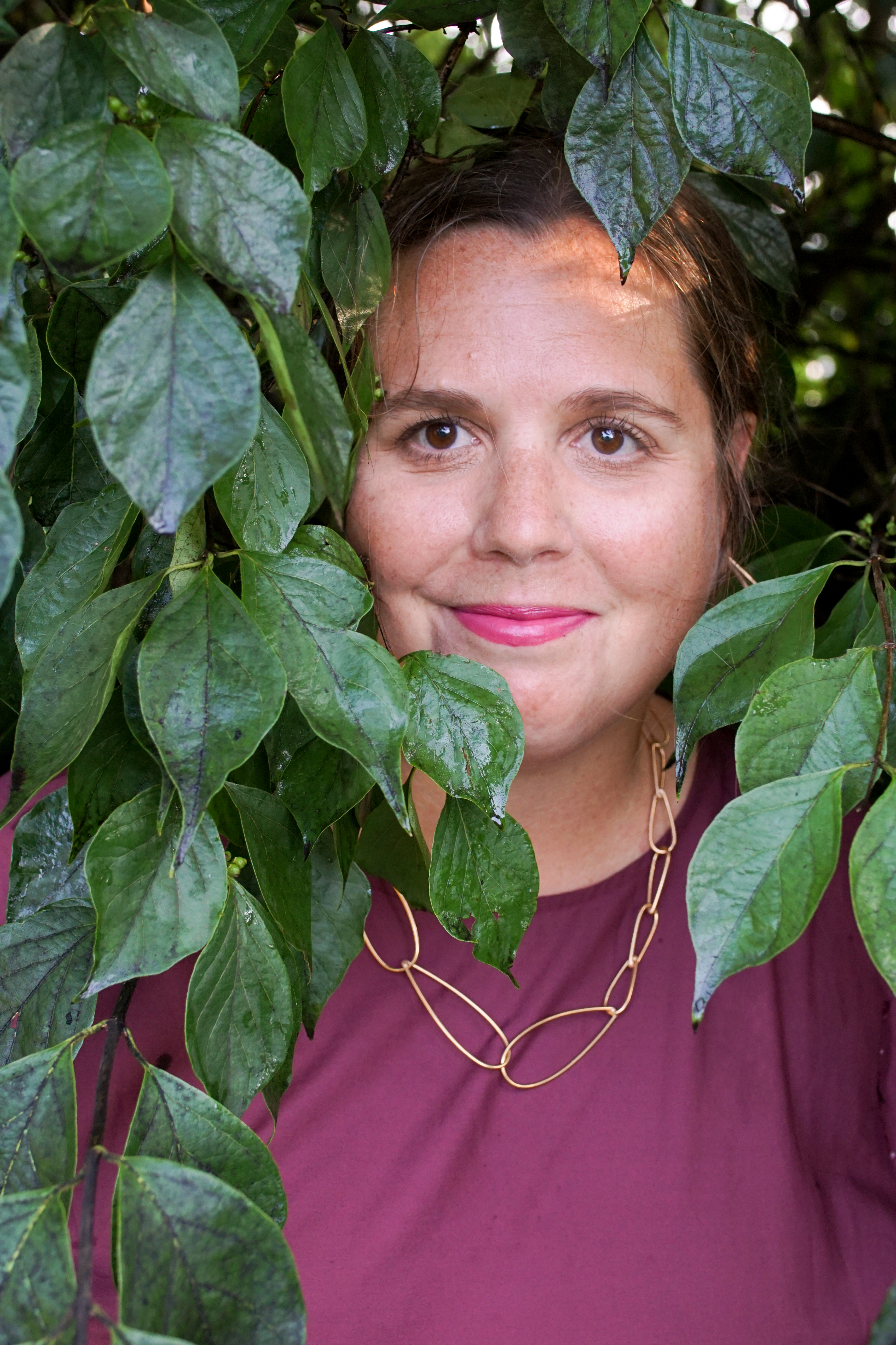 woman wearing burgundy dress and bronze chain necklace standing with a bush