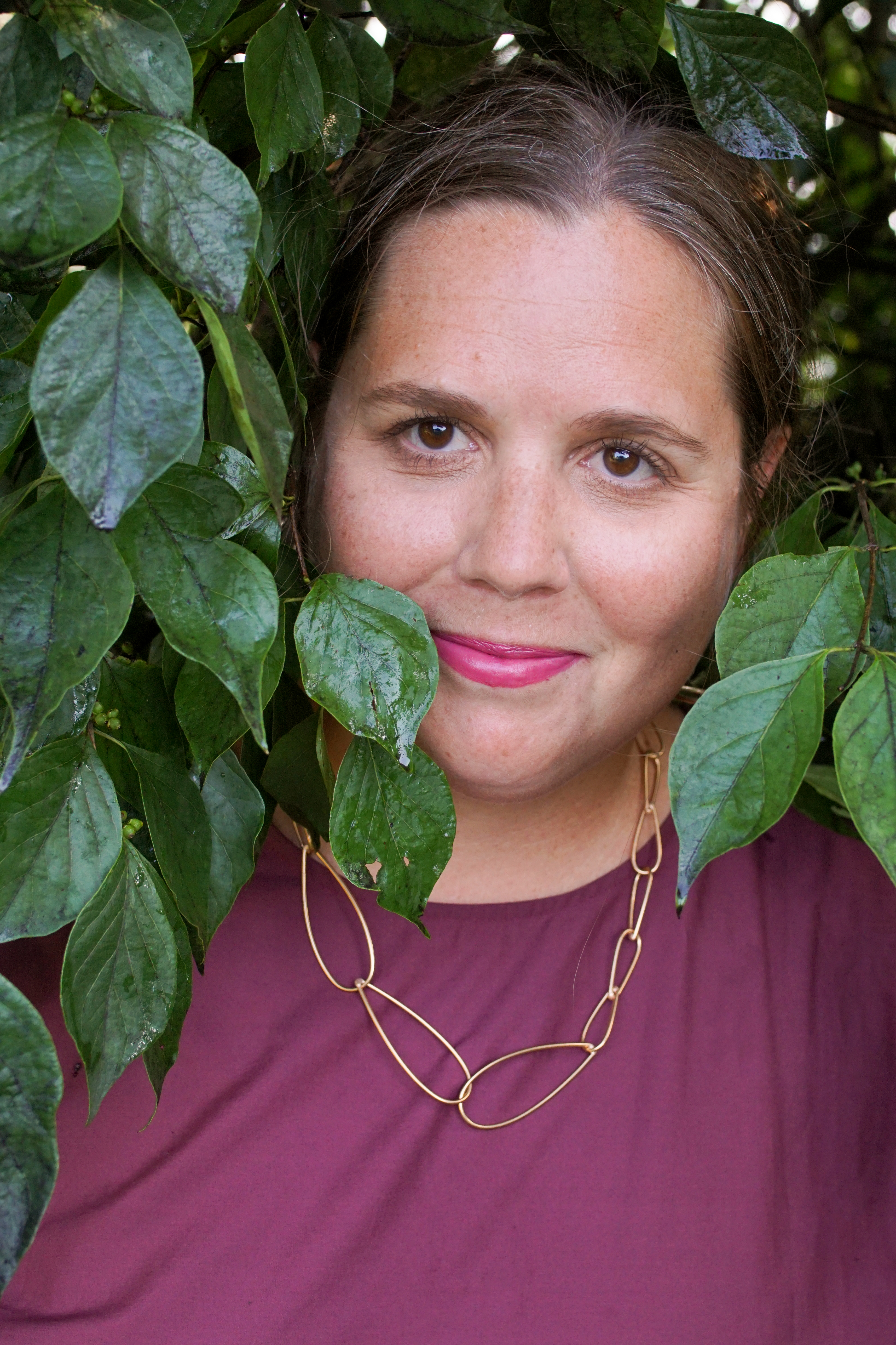 woman wearing burgundy dress and bronze chain necklace standing with a bush