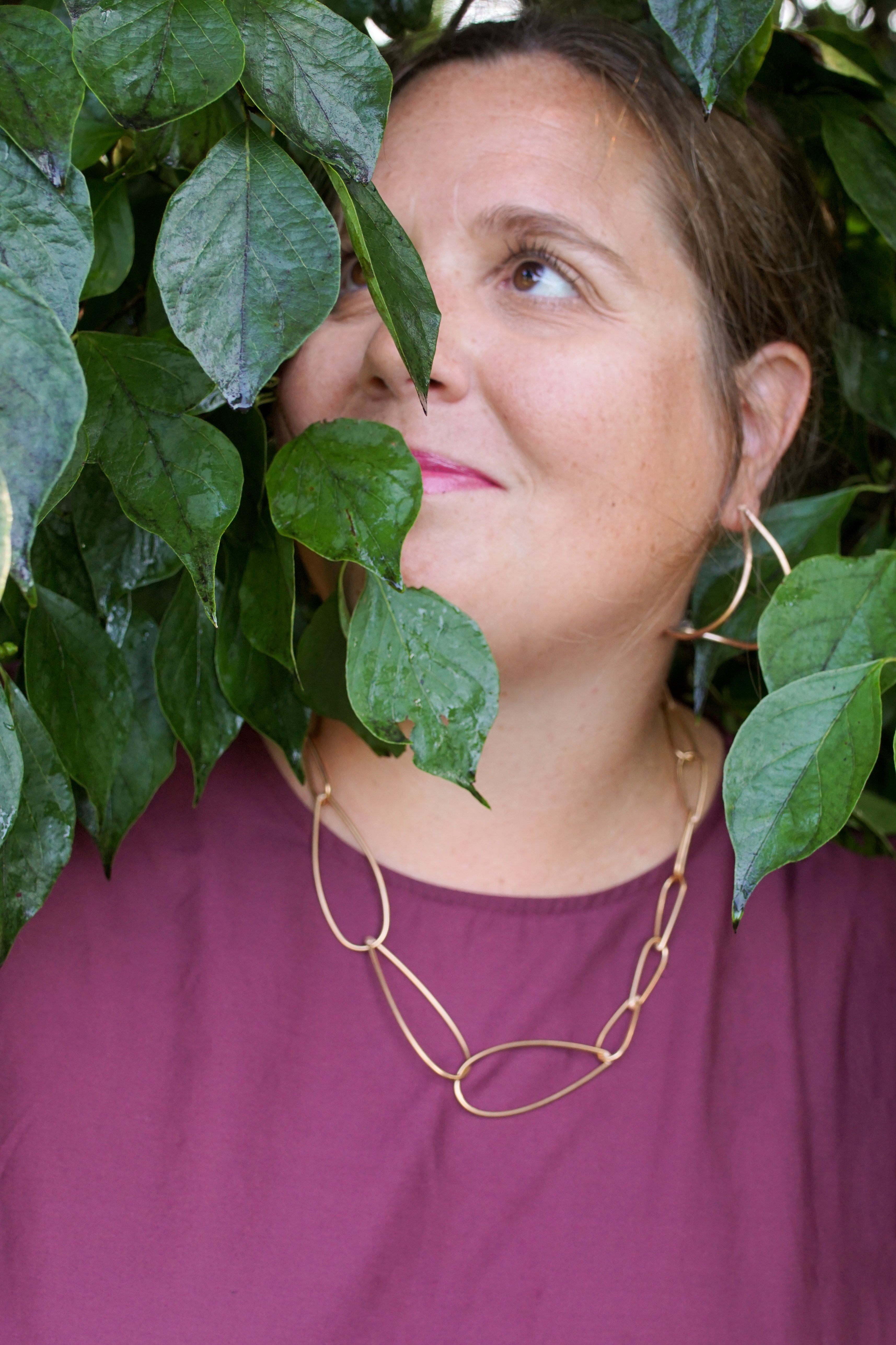 woman wearing burgundy dress and bronze chain necklace standing with a bush