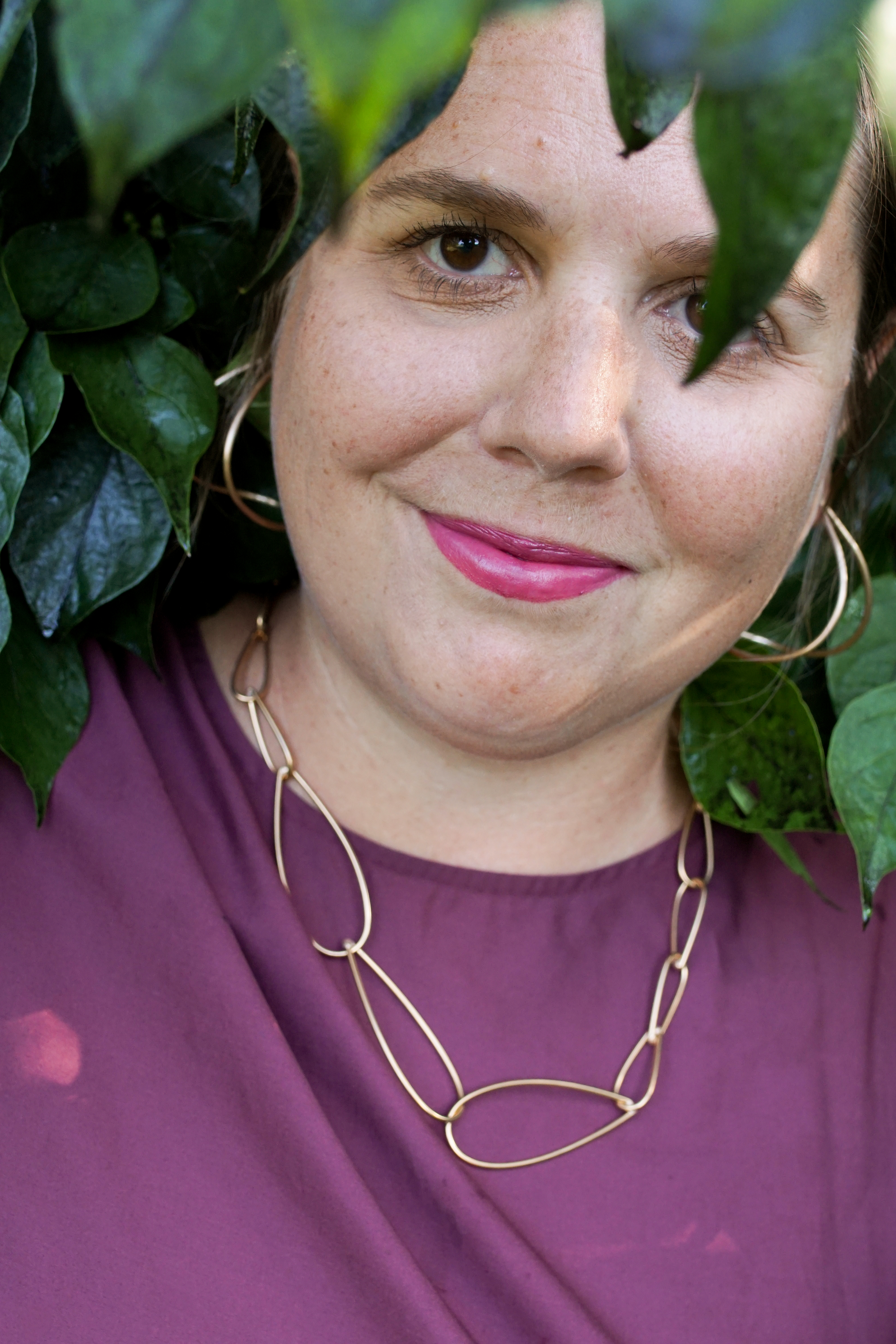 woman wearing burgundy dress and bronze chain necklace standing with a bush