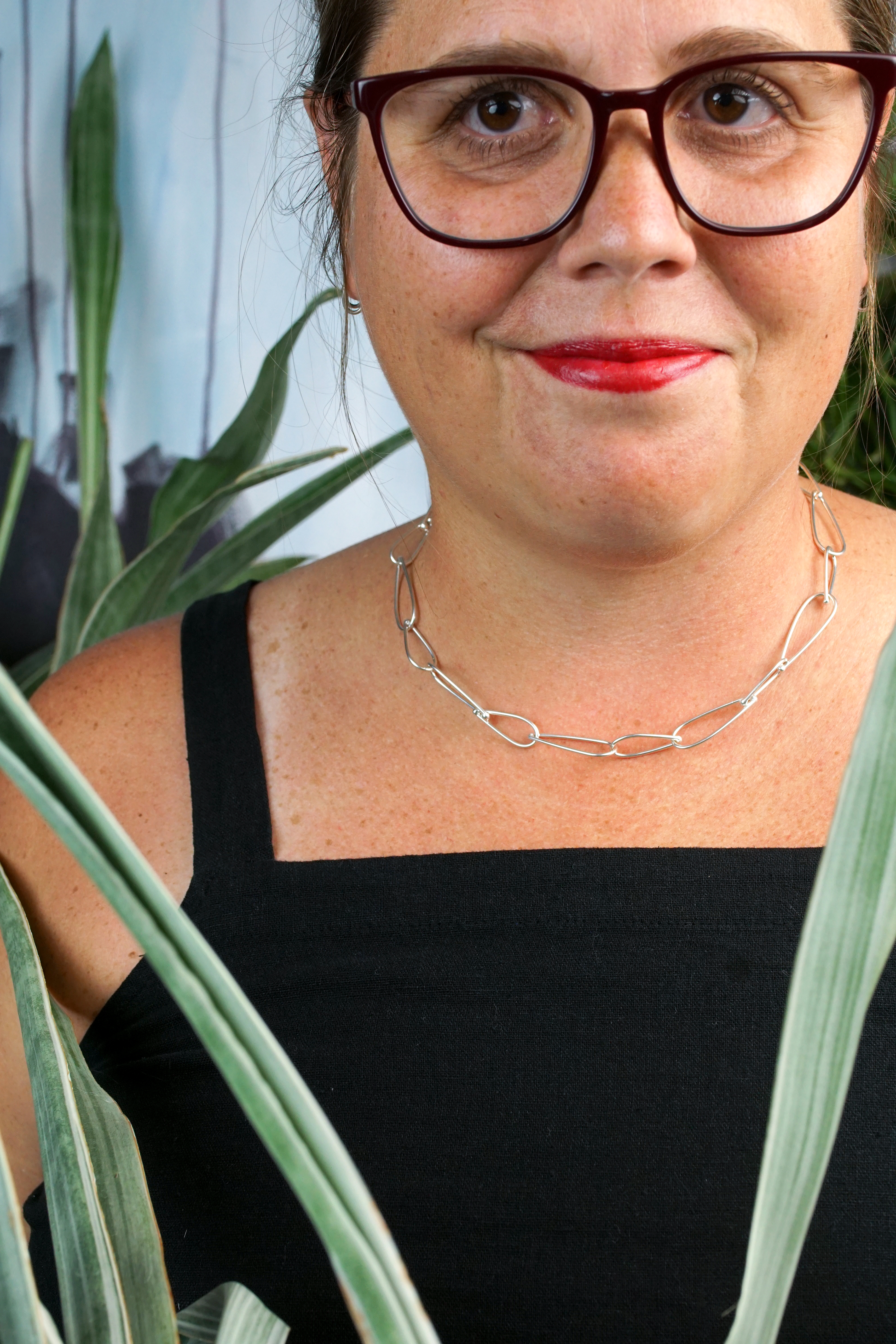 french librarian vibes: woman wearing black jumpsuit, silver chain necklace, glasses and red lipstick posing with plants