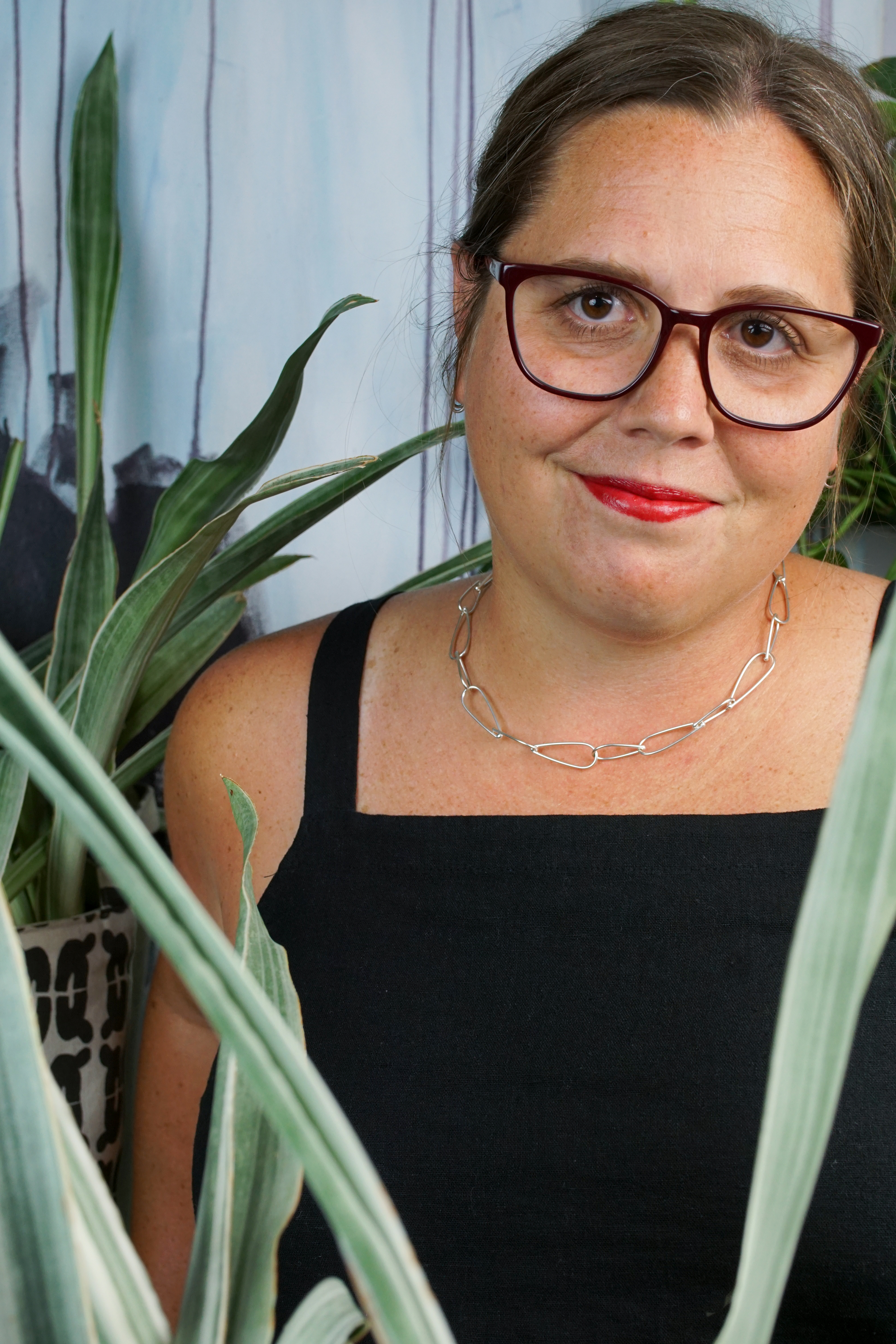 french librarian vibes: woman wearing black jumpsuit, silver chain necklace, glasses and red lipstick posing with plants