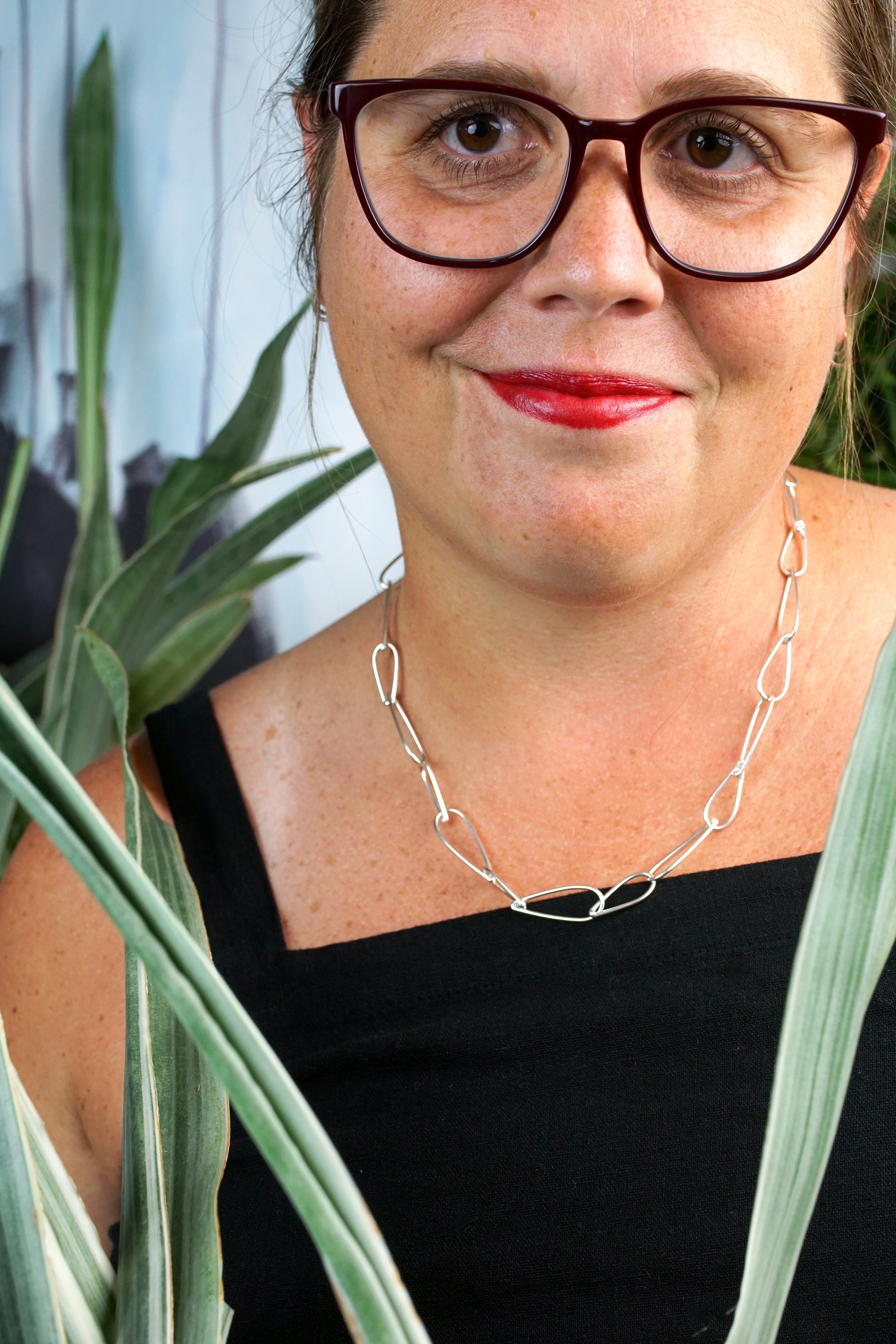 french librarian vibes: woman wearing black jumpsuit, silver chain necklace, glasses and red lipstick posing with plants