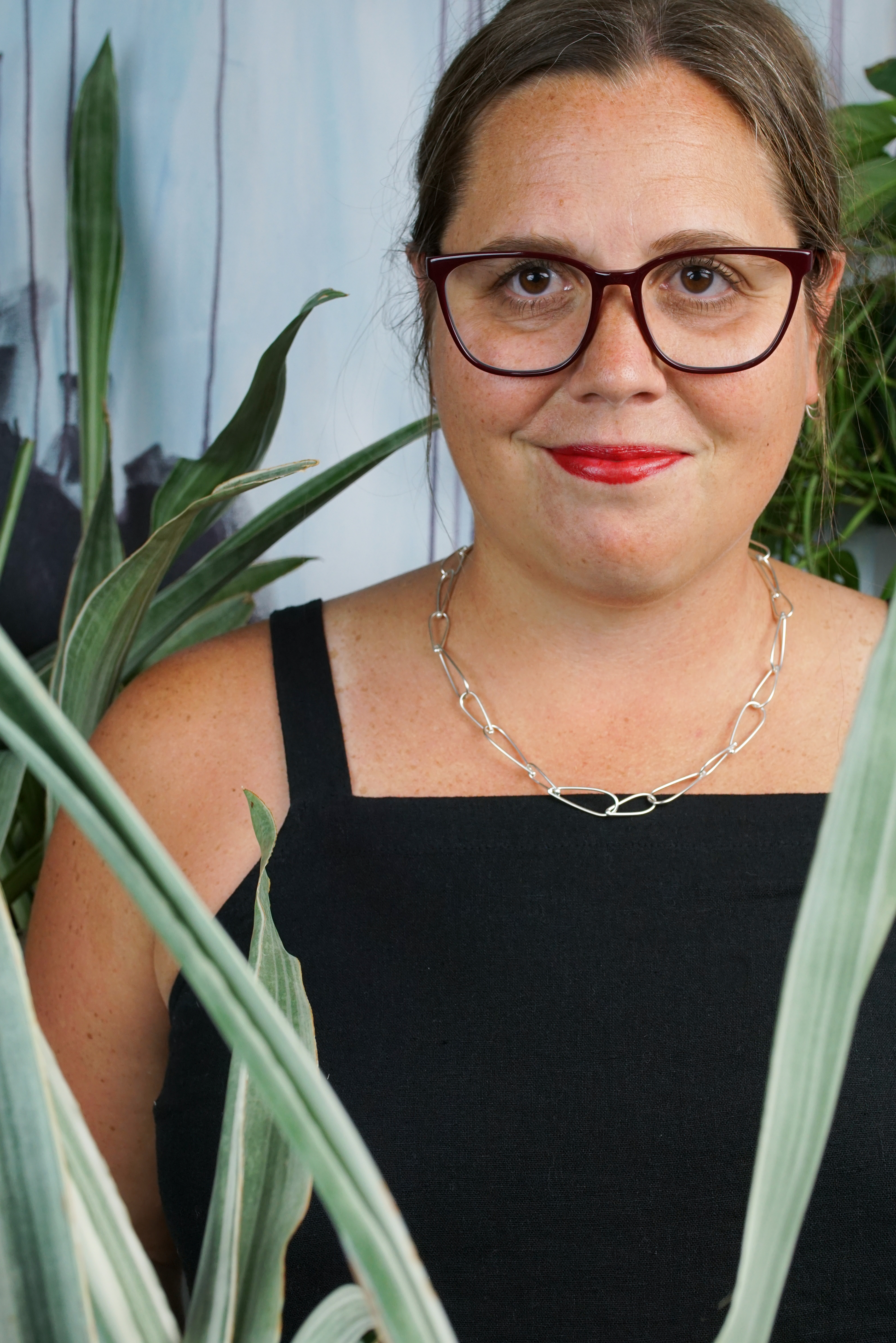 french librarian vibes: woman wearing black jumpsuit, silver chain necklace, glasses and red lipstick posing with plants