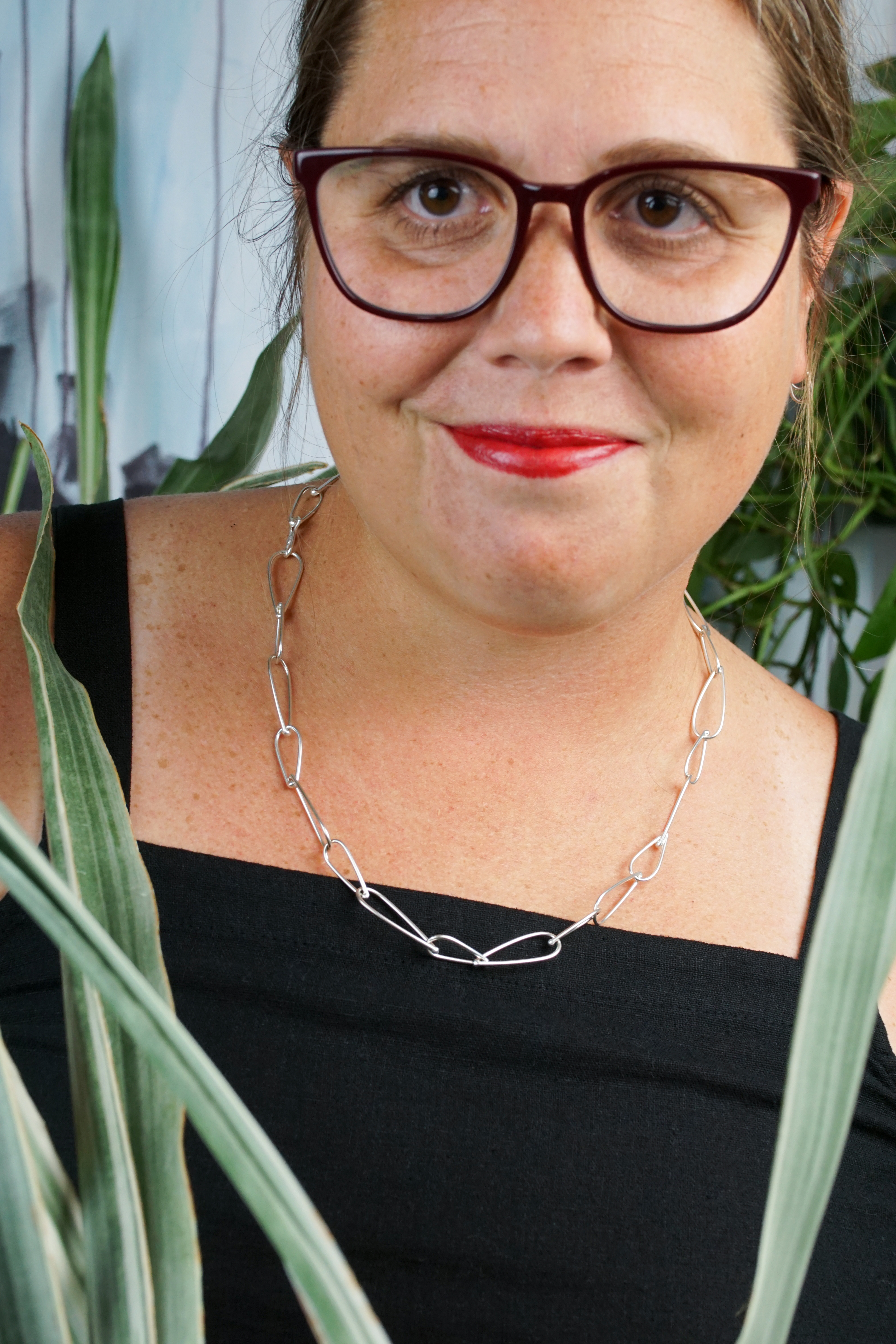 french librarian vibes: woman wearing black jumpsuit, silver chain necklace, glasses and red lipstick posing with plants