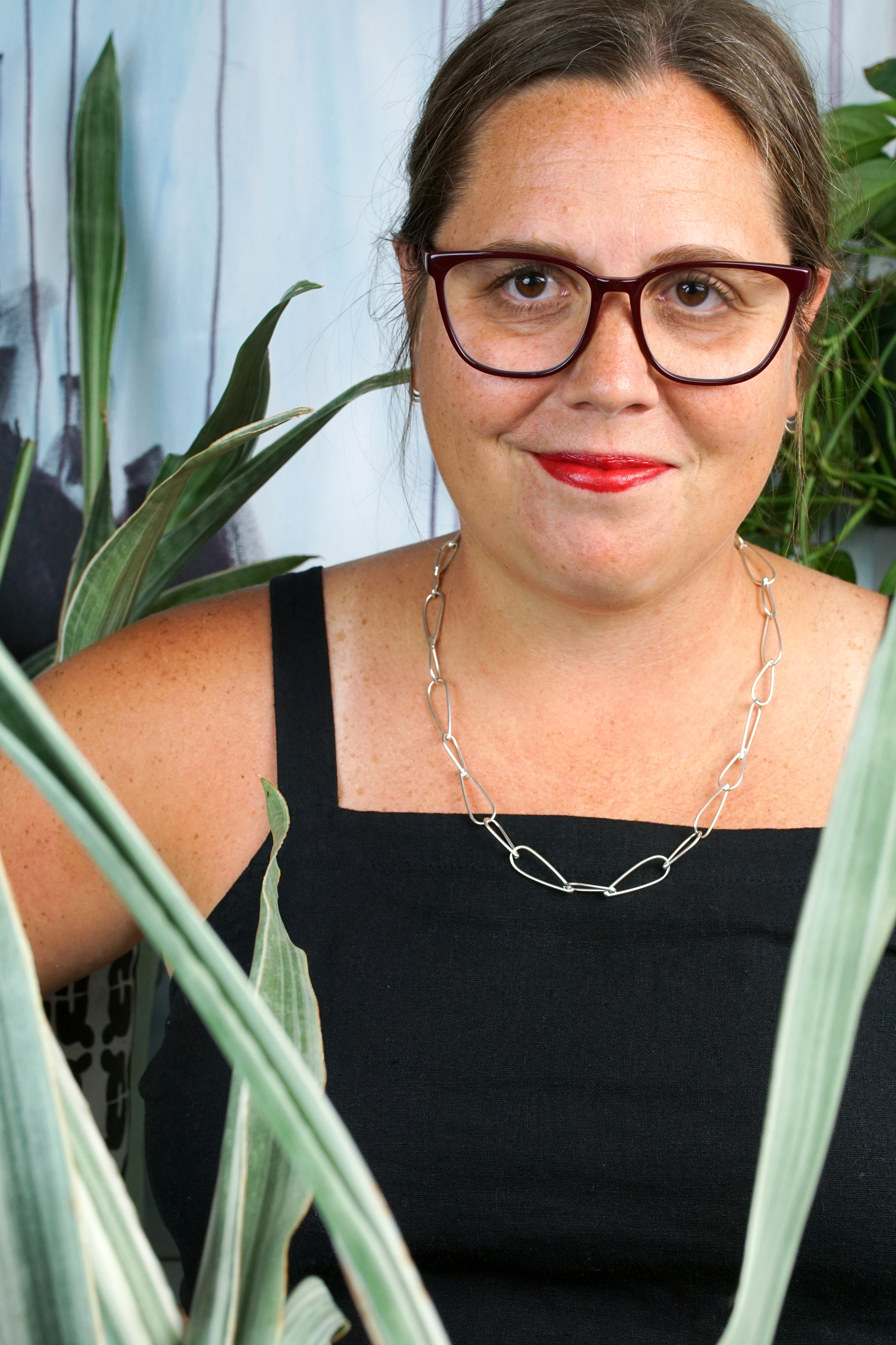 french librarian vibes: woman wearing black jumpsuit, silver chain necklace, glasses and red lipstick posing with plants