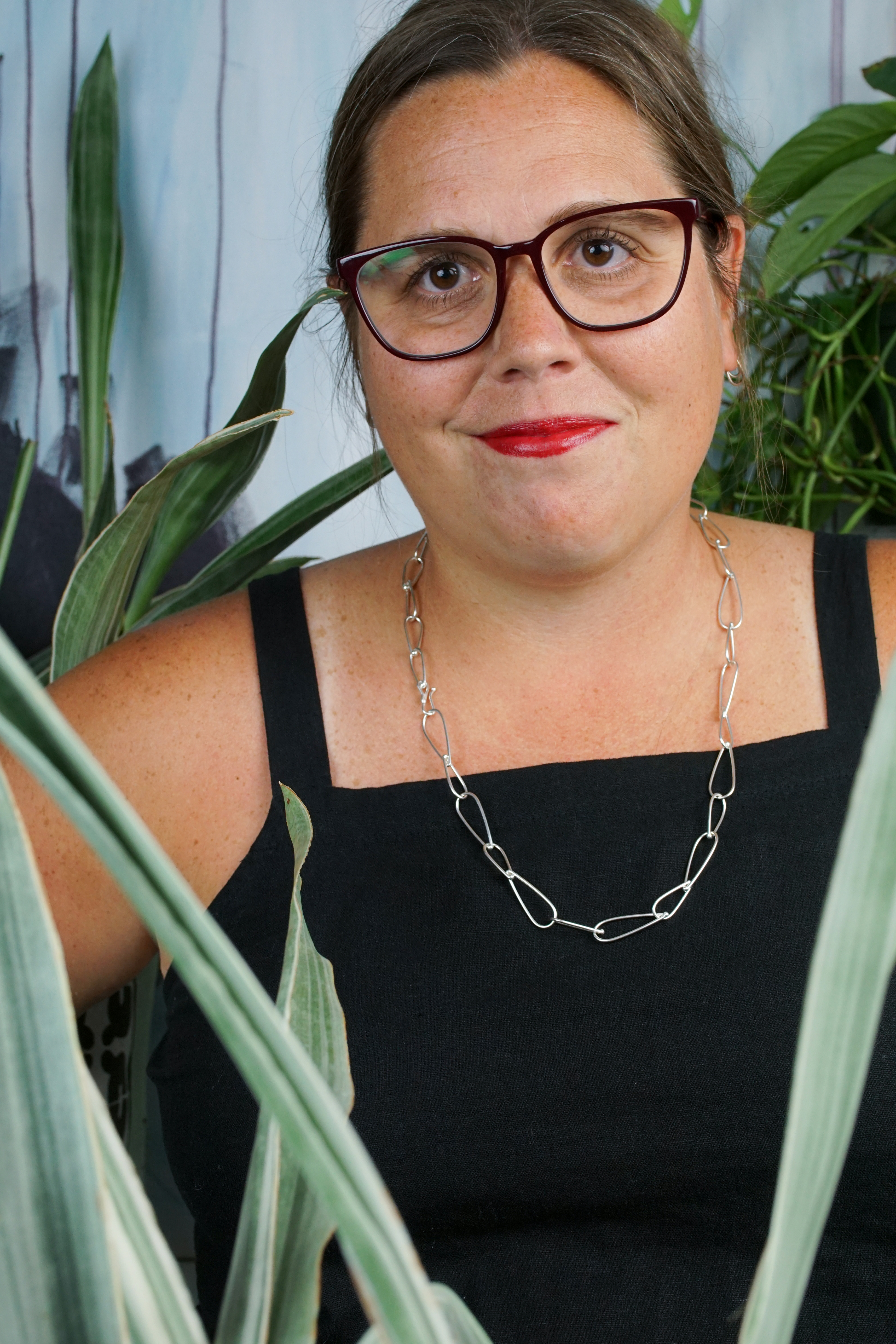 french librarian vibes: woman wearing black jumpsuit, silver chain necklace, glasses and red lipstick posing with plants