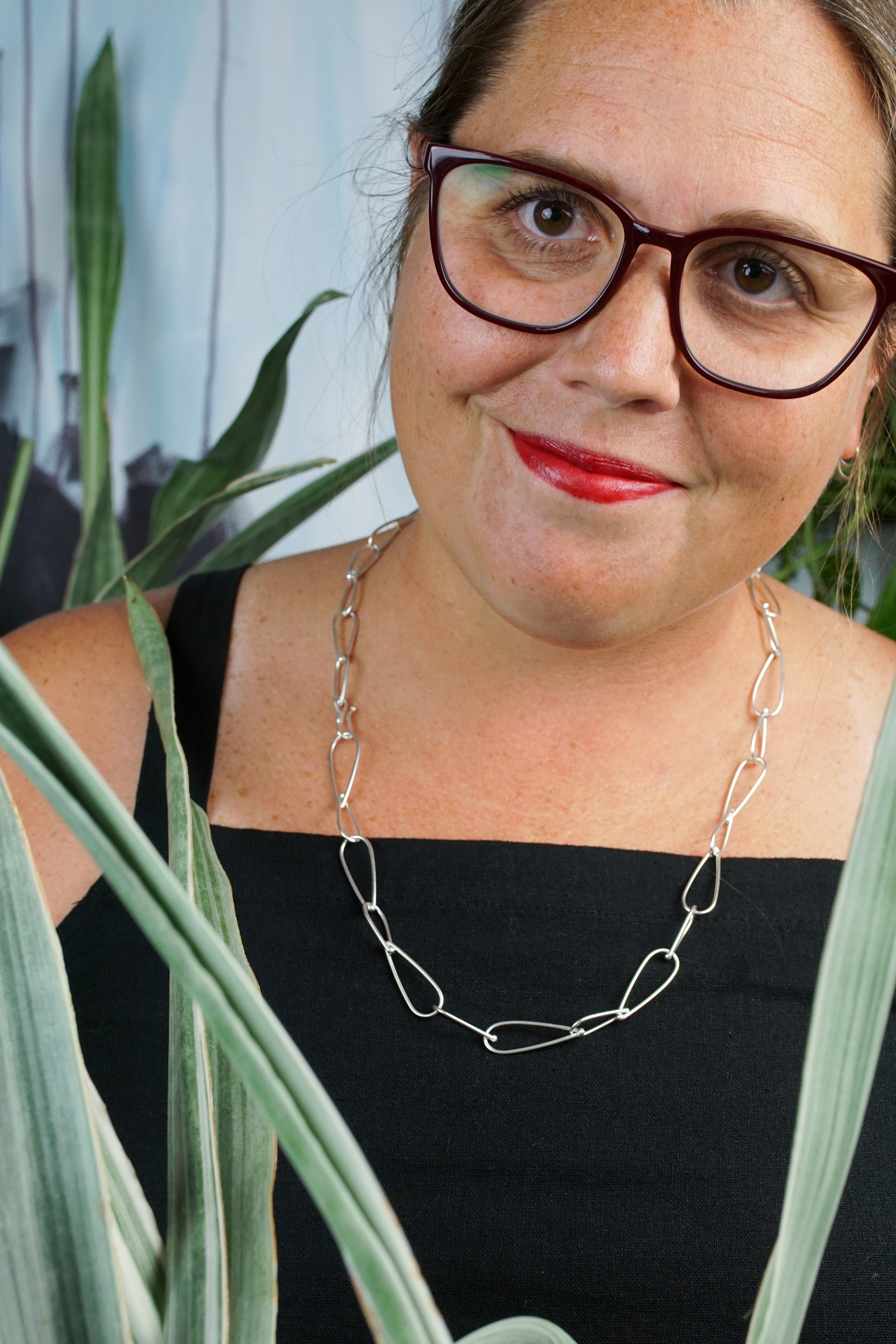 french librarian vibes: woman wearing black jumpsuit, silver chain necklace, glasses and red lipstick posing with plants