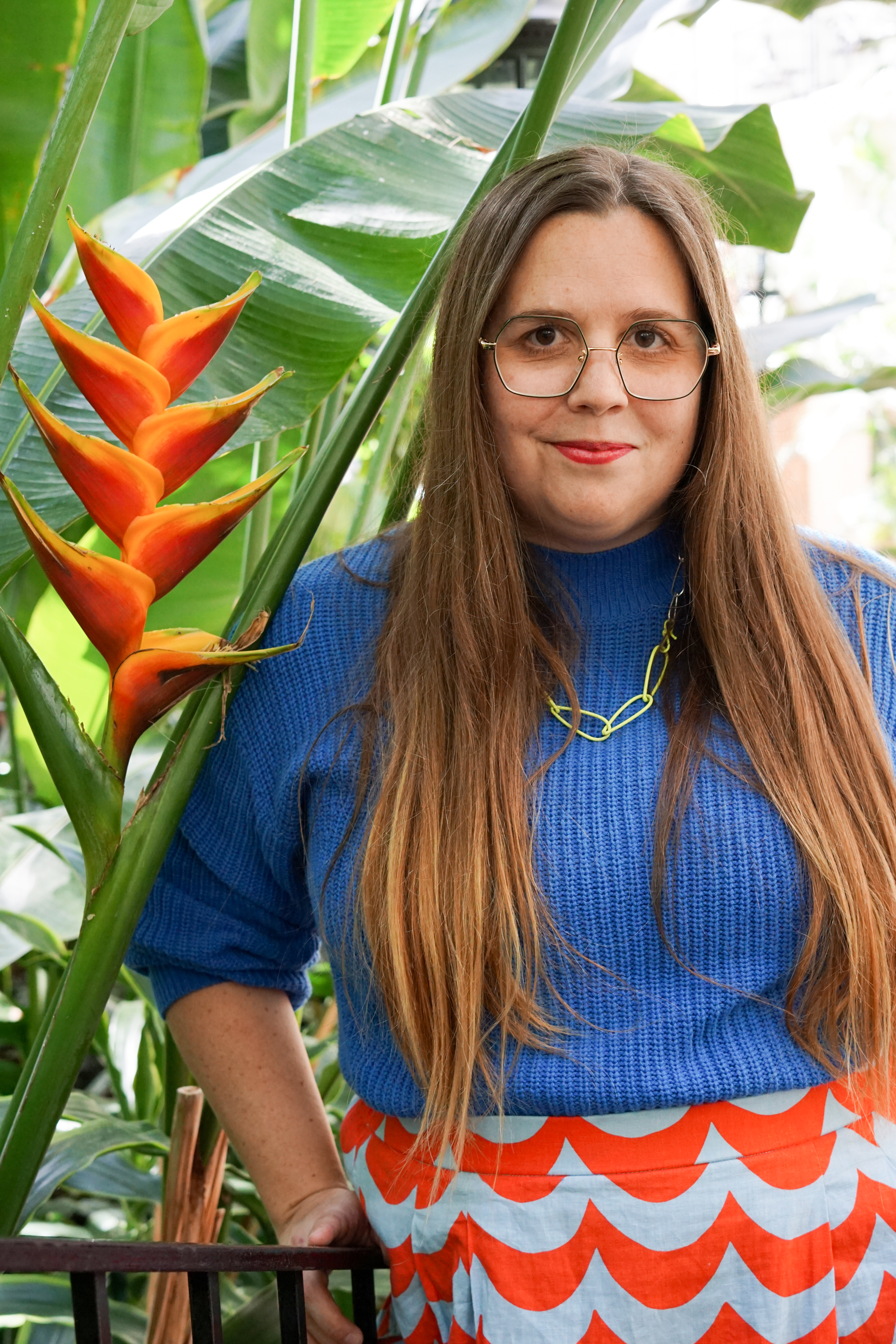 self-portrait in garden with bird of paradise plant, neon necklace, blue sweater, and marimekko skirt