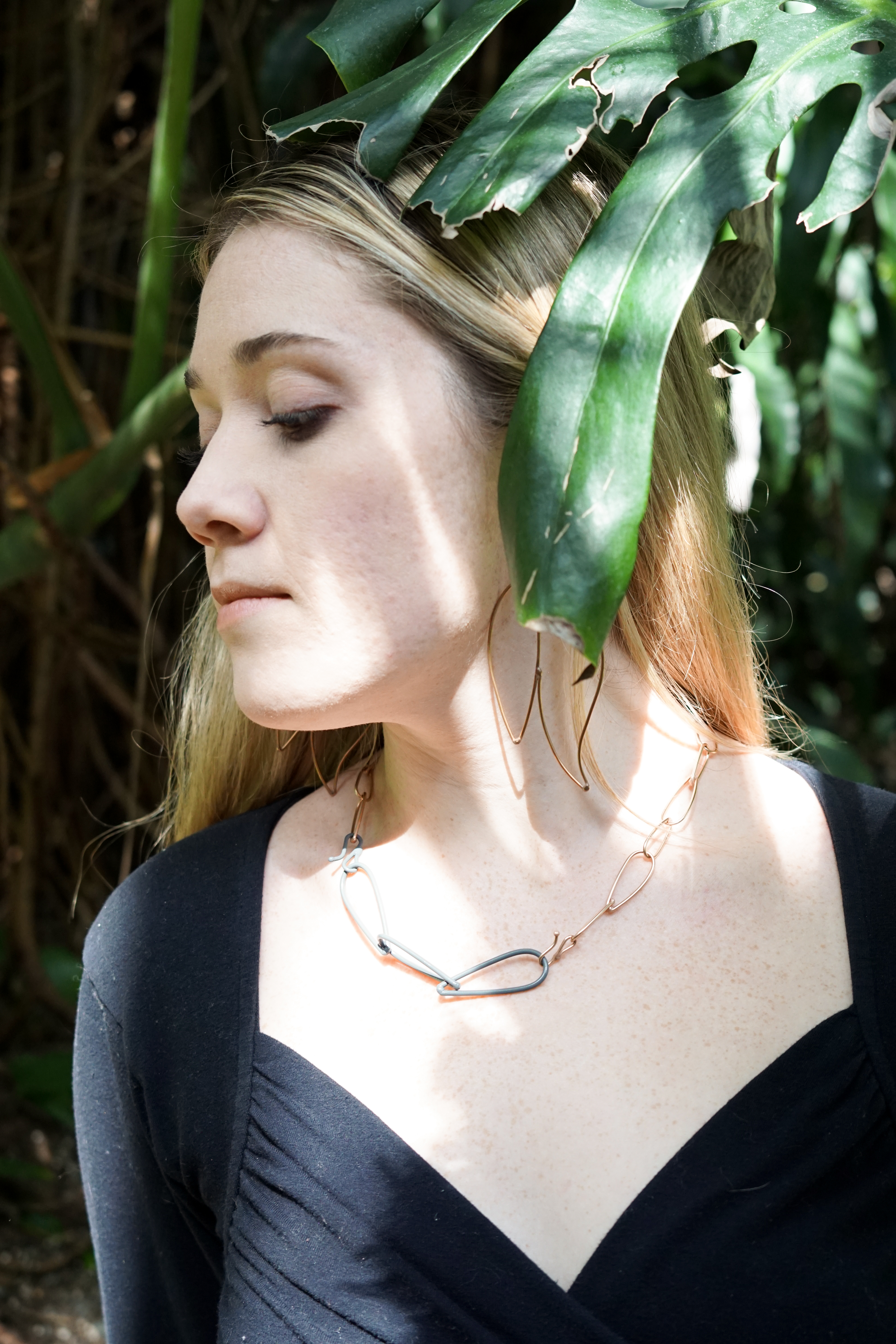 Trista in the garden with a monstera leaf wearing bronze statement earrings and a bronze and grey chain link necklace