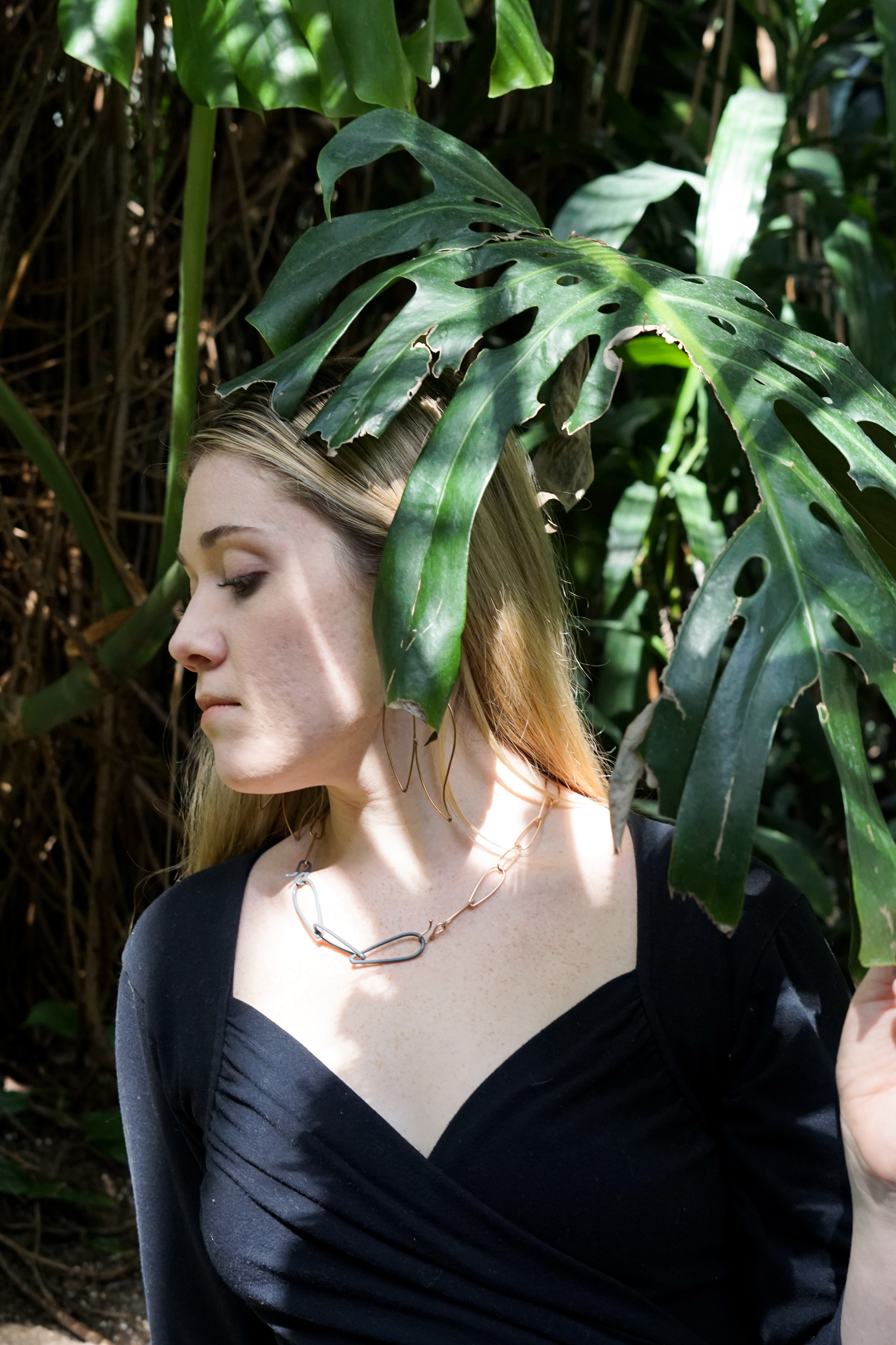 Trista in the garden with a monstera leaf wearing bronze statement earrings and a bronze and grey chain link necklace