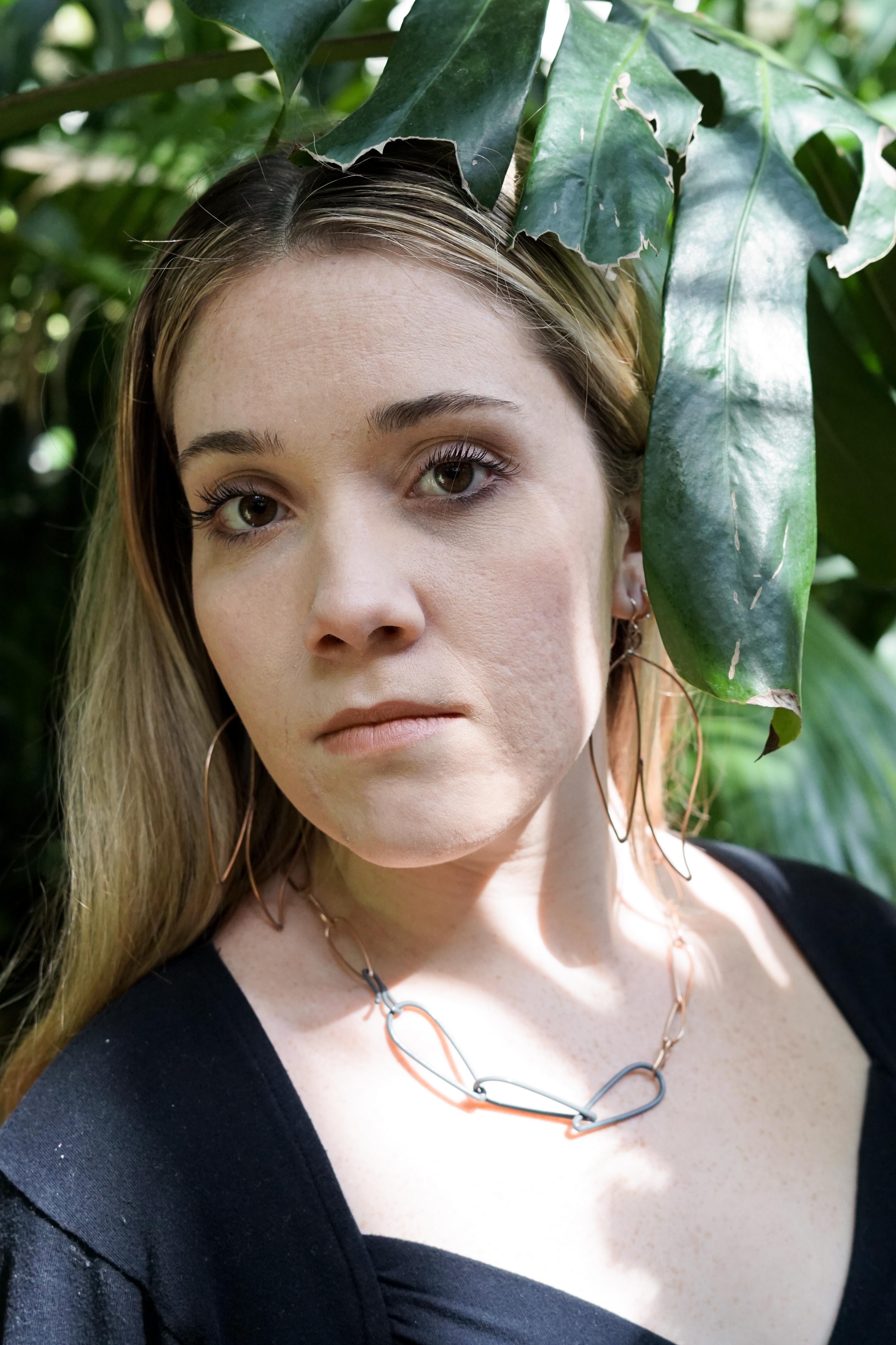 Trista in the garden with a monstera leaf wearing bronze statement earrings and a bronze and grey chain link necklace