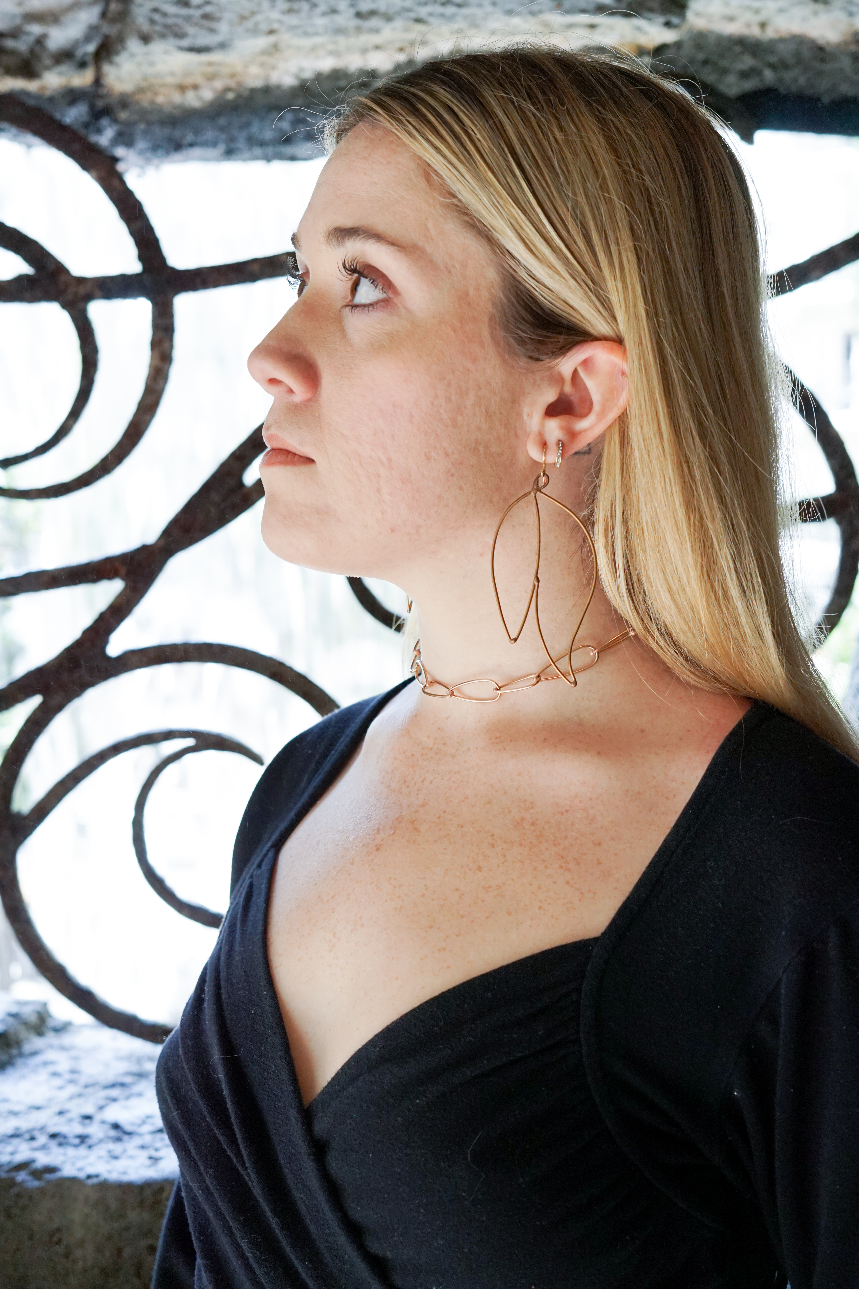 Trista wearing bronze statement earrings and bronze chain necklace standing in front of decorative ironwork