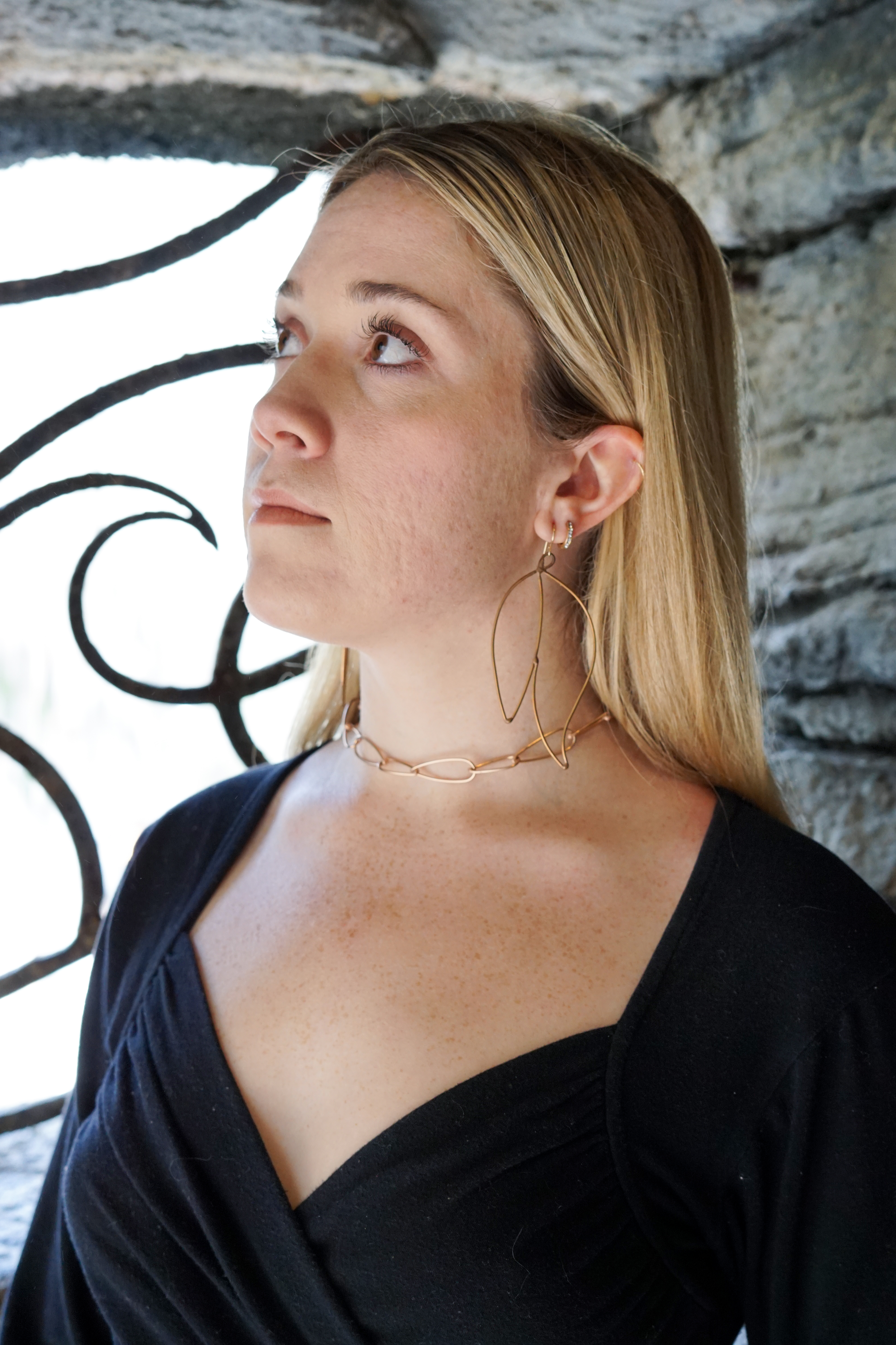 Trista wearing bronze statement earrings and bronze chain necklace standing in front of decorative ironwork