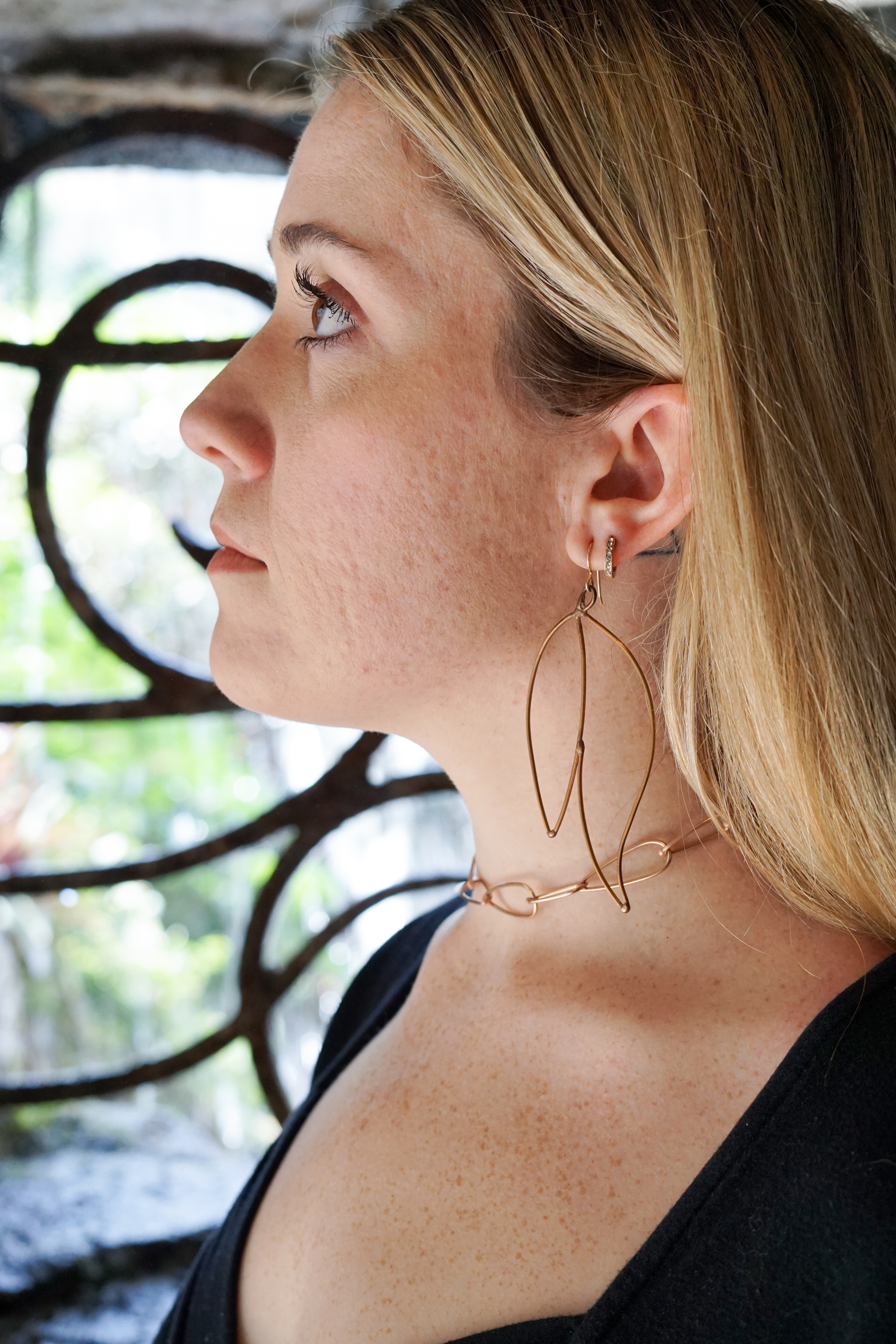 Trista wearing bronze statement earrings and bronze chain necklace standing in front of decorative ironwork